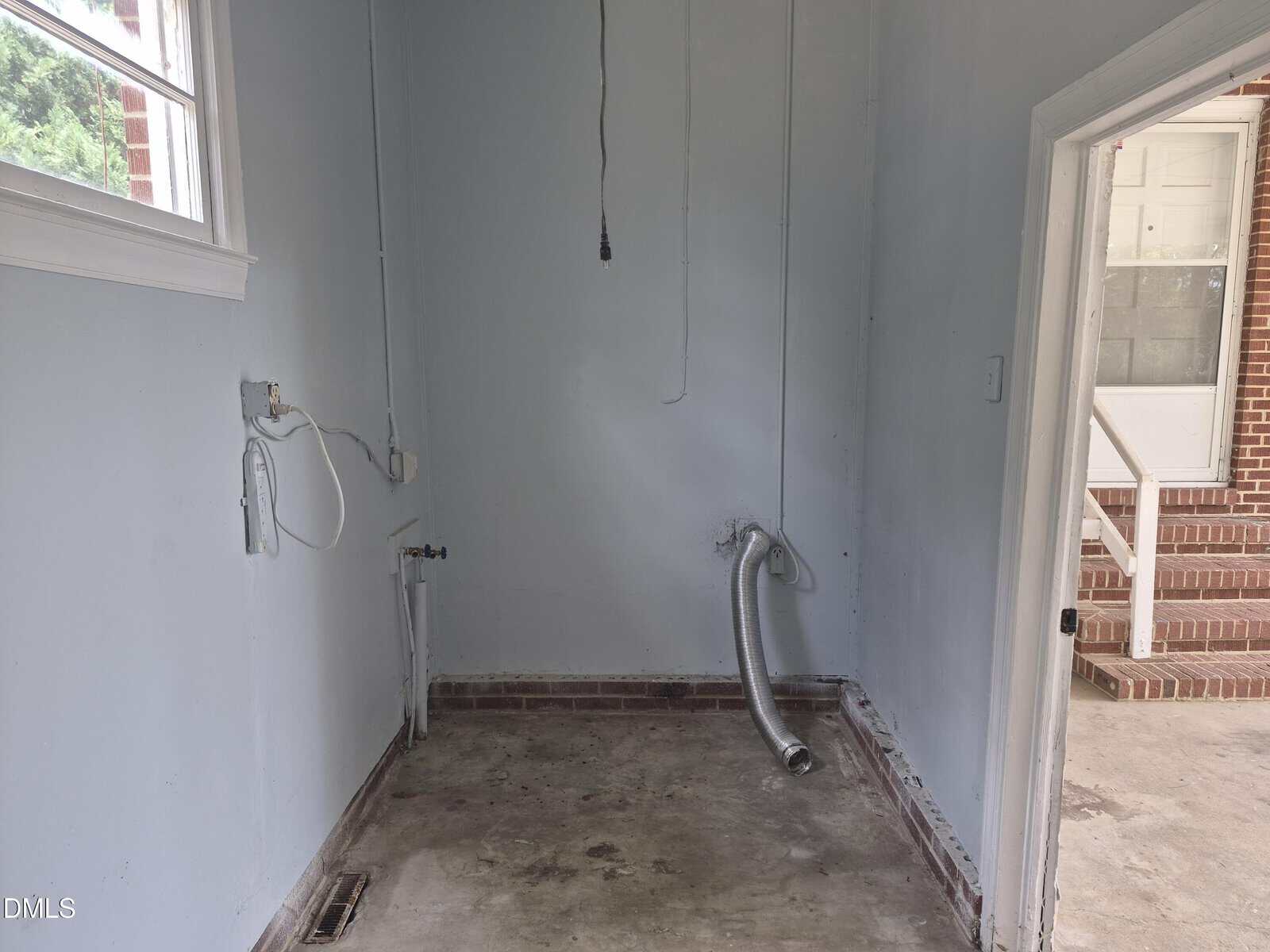 Empty laundry room with exposed dryer vent hose, utility hookups, and a concrete floor. A window and open door reveal the outside brick steps and another door. Walls are painted light blue.