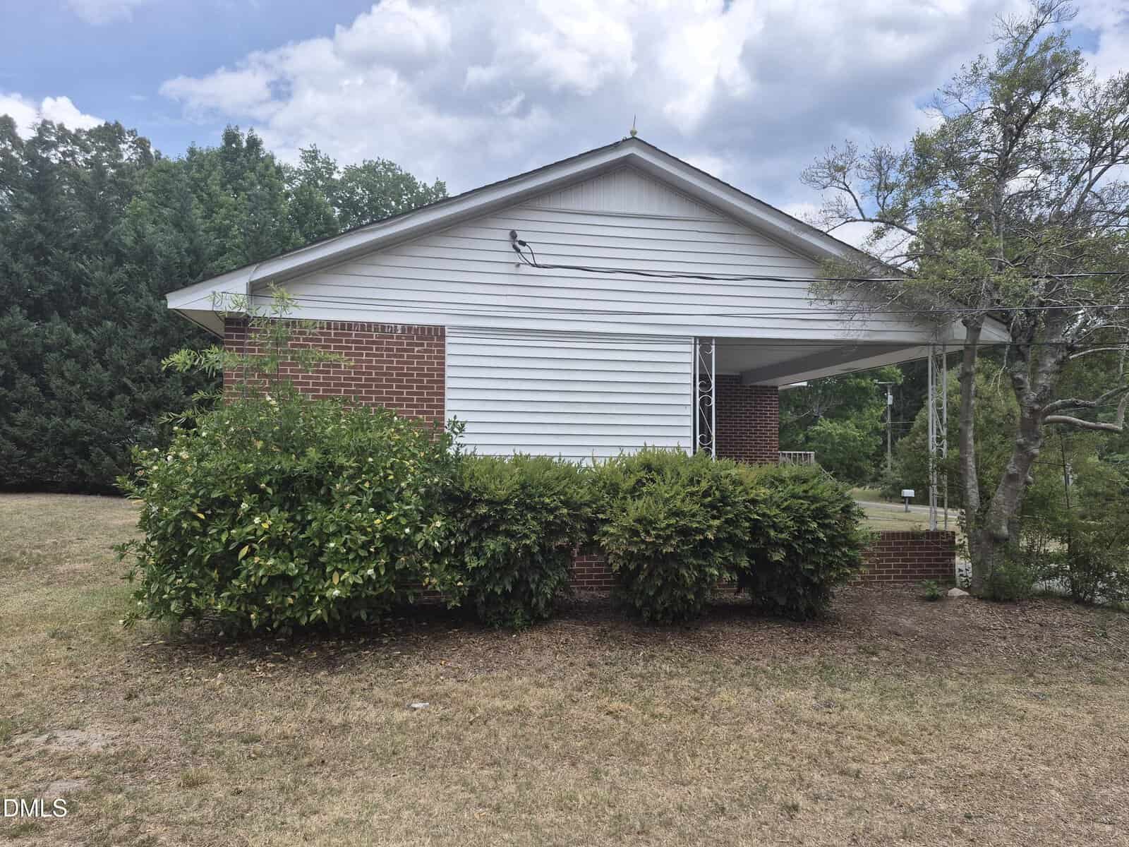 Side view of a one-story house with red brick and white siding, shrubs in front, a sloped roof, and a cloudy sky overhead. The yard is dry with patches of brown grass. Trees are in the background.
