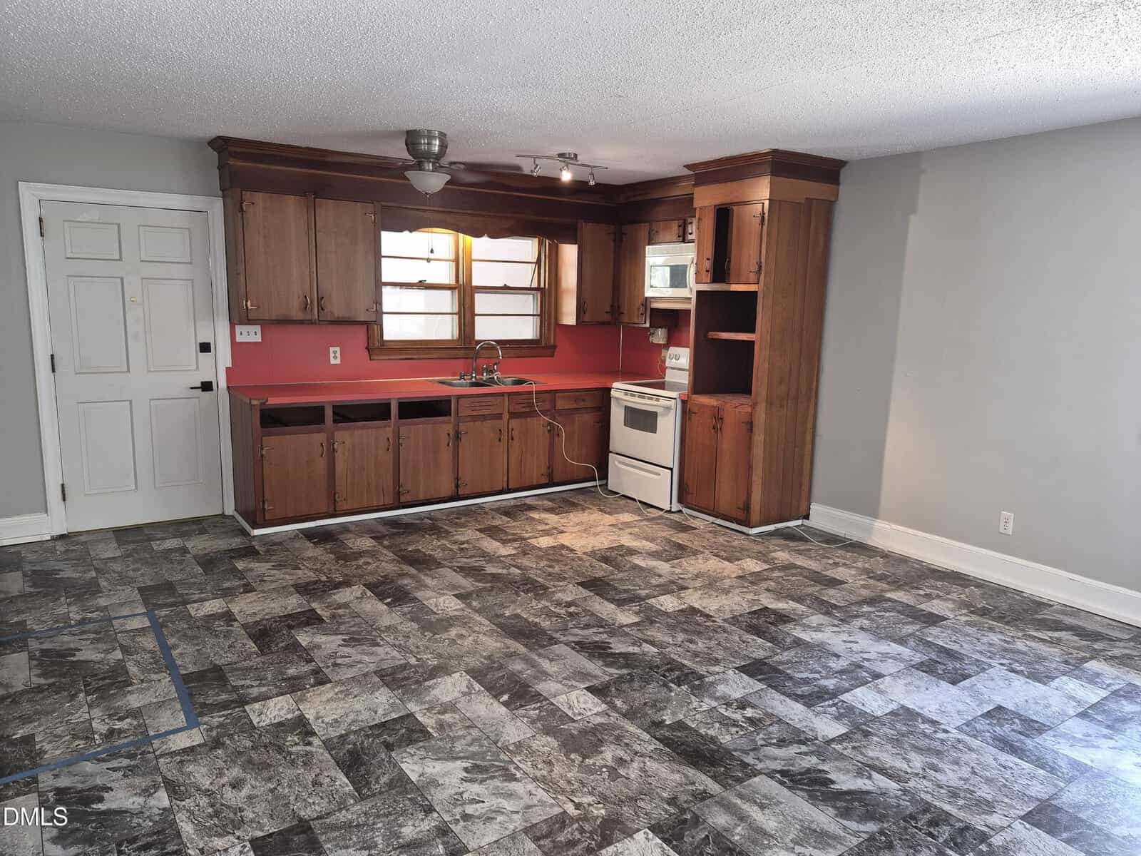 Small kitchen with wooden cabinets, a white stove, and a stainless steel sink under a window. The backsplash is painted red. The room has gray patterned flooring and light gray walls, with a white door on the left.