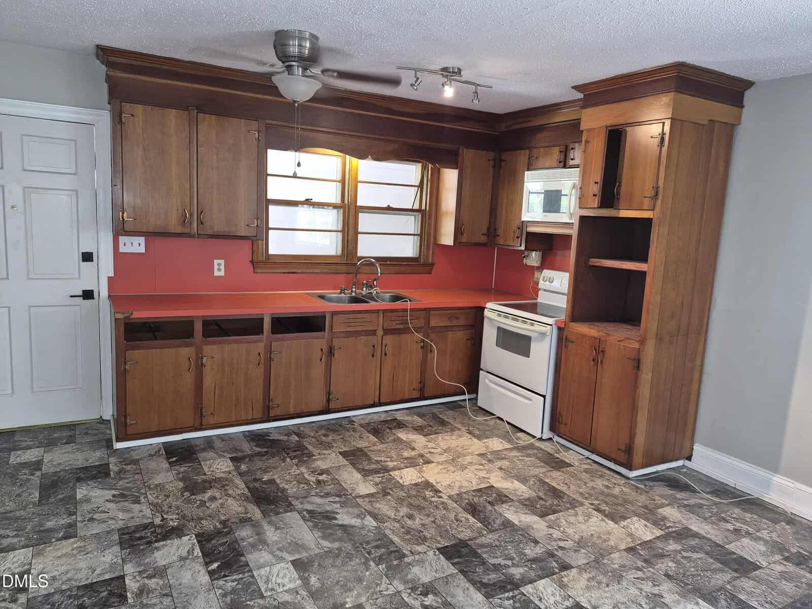A small kitchen with wooden cabinets missing doors and drawers, a white stove, red backsplash, double sink under two windows, and gray patterned floor tiles. A ceiling fan and light fixture are installed above.