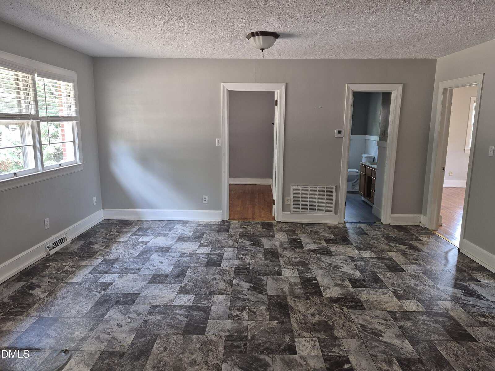 A mostly empty room with a patterned tile floor, gray walls, a ceiling light, and three doorways leading to other rooms. Sunlight comes through windows on the left. A bathroom is visible through one doorway.
