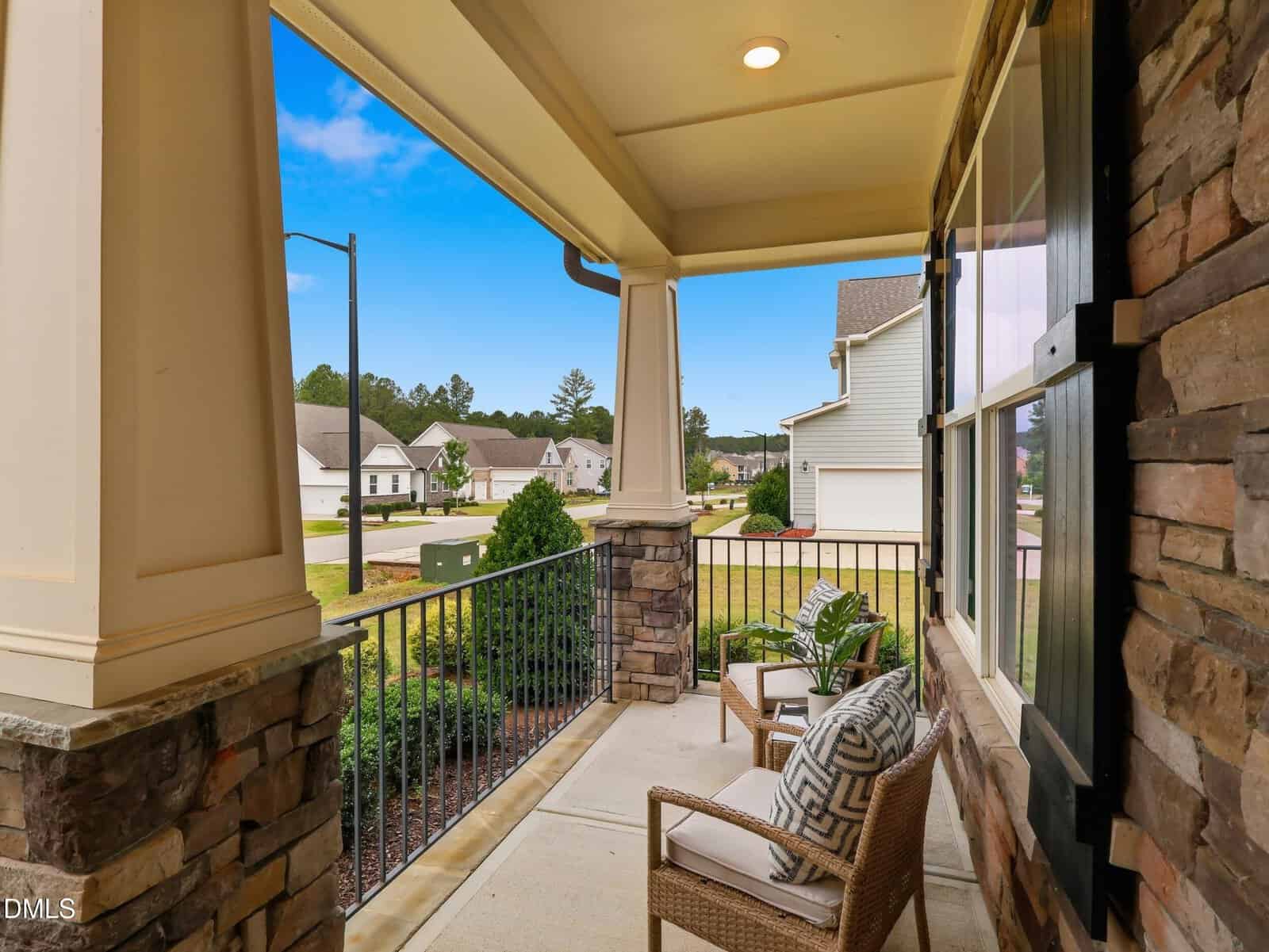 A covered front porch with two cushioned chairs, stone pillars, and a view of a suburban neighborhood with green lawns and neighboring houses under a partly cloudy sky.