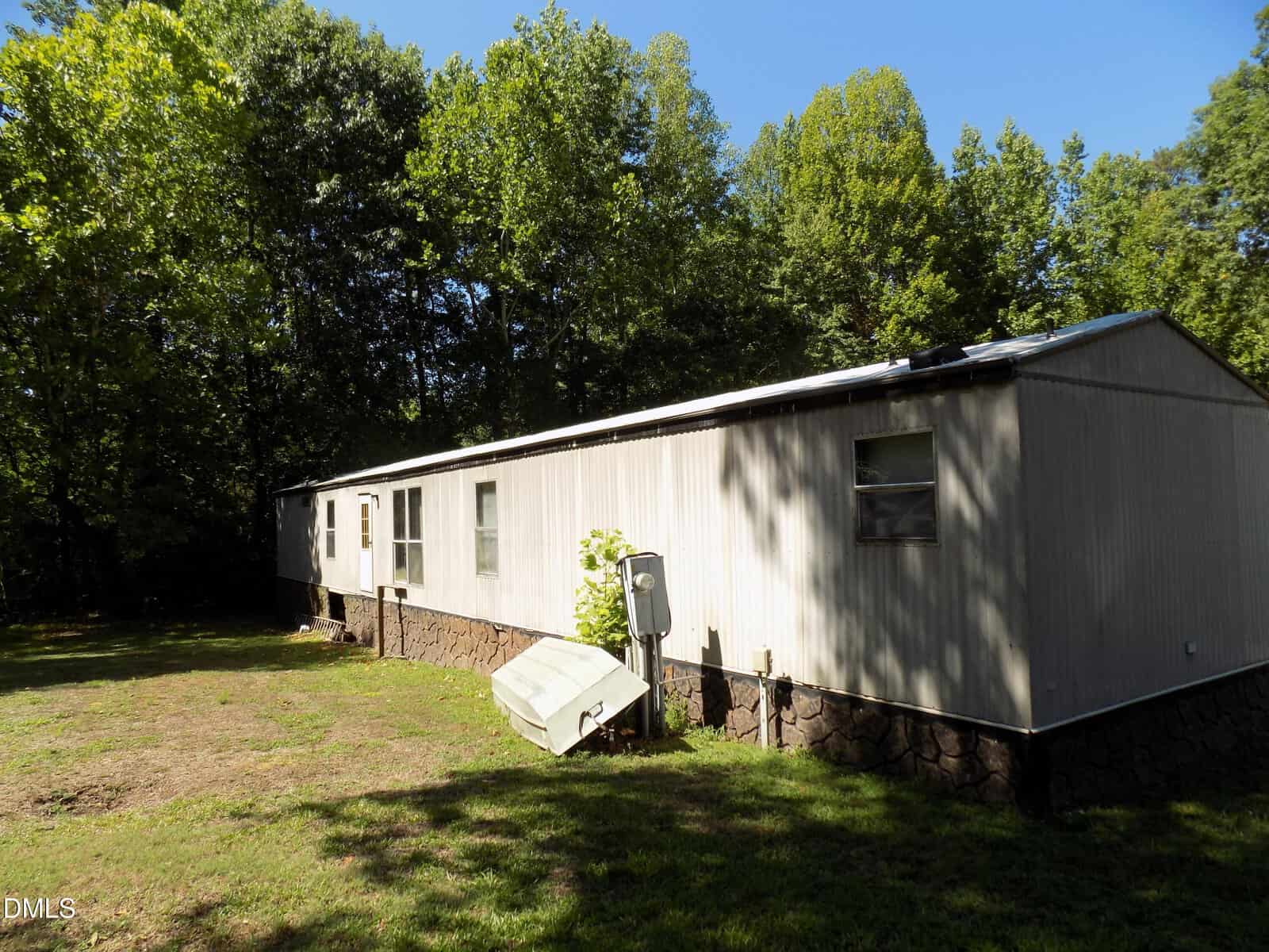A long, beige mobile home with a stone foundation sits on a grassy yard, surrounded by tall green trees under a clear blue sky.