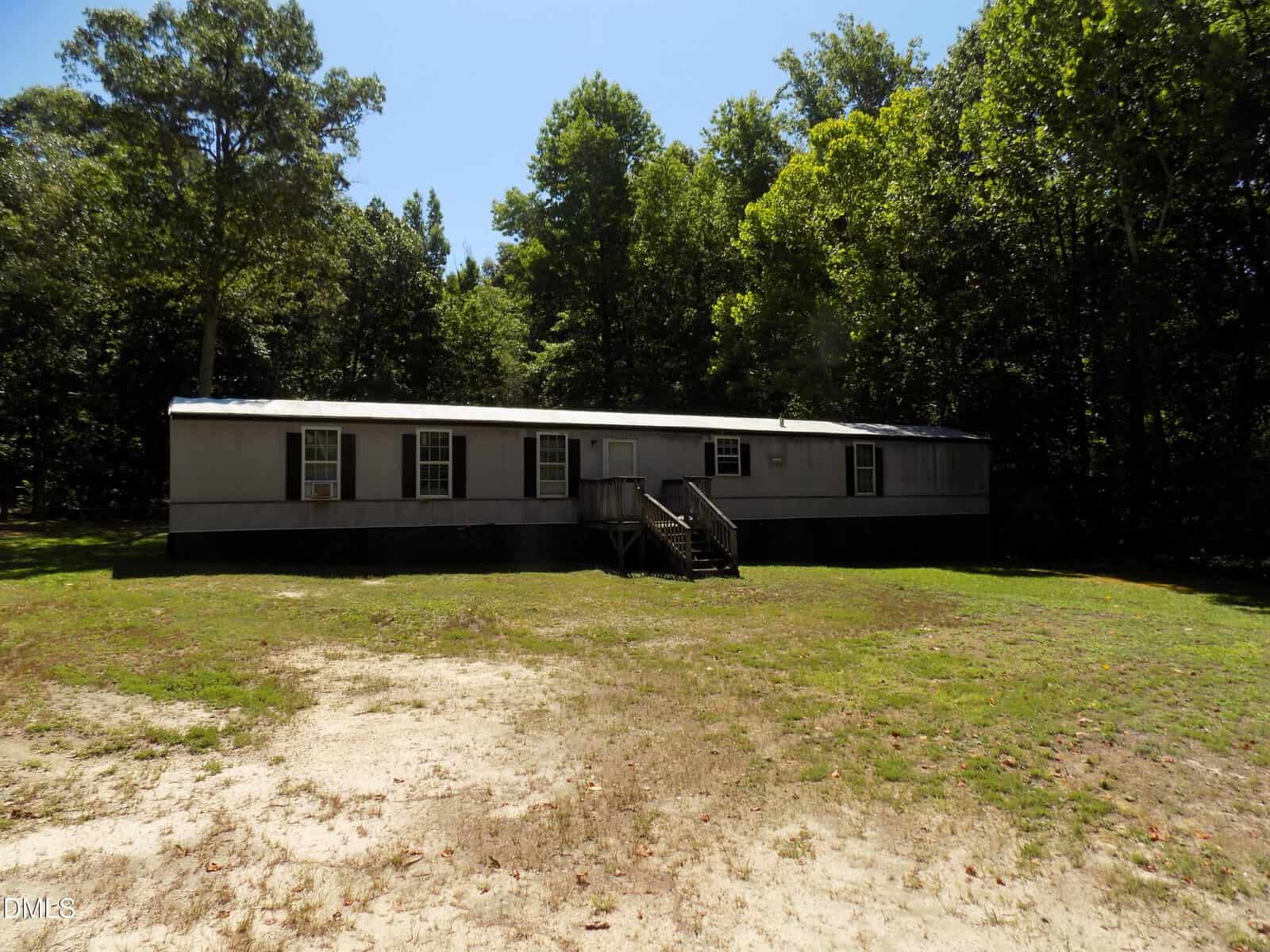 A single-story mobile home with light-colored siding and multiple windows sits on a grassy, partially shaded lot surrounded by tall trees under a clear blue sky. Simple steps lead up to the front door.
