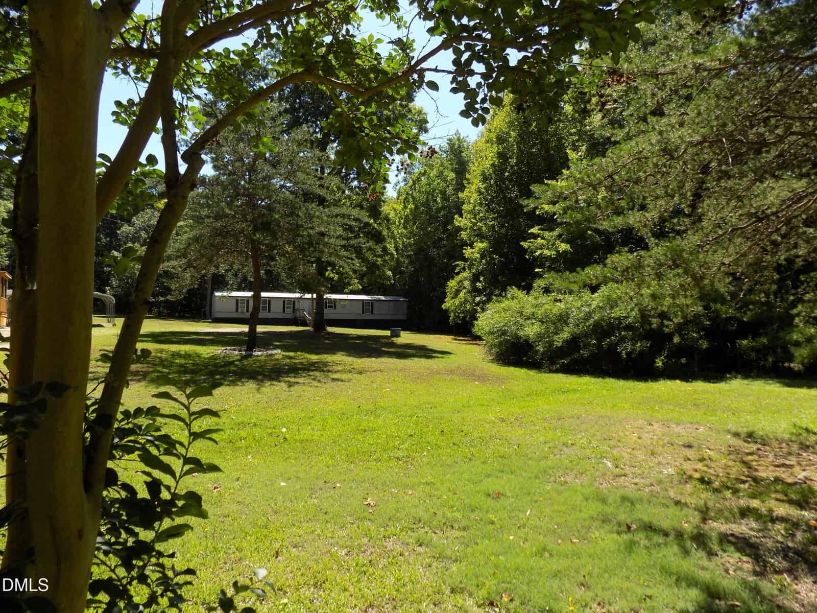 A single-story white mobile home sits in the distance, surrounded by lush green grass, trees, and dense forest on a sunny day, with shadows cast on the lawn.