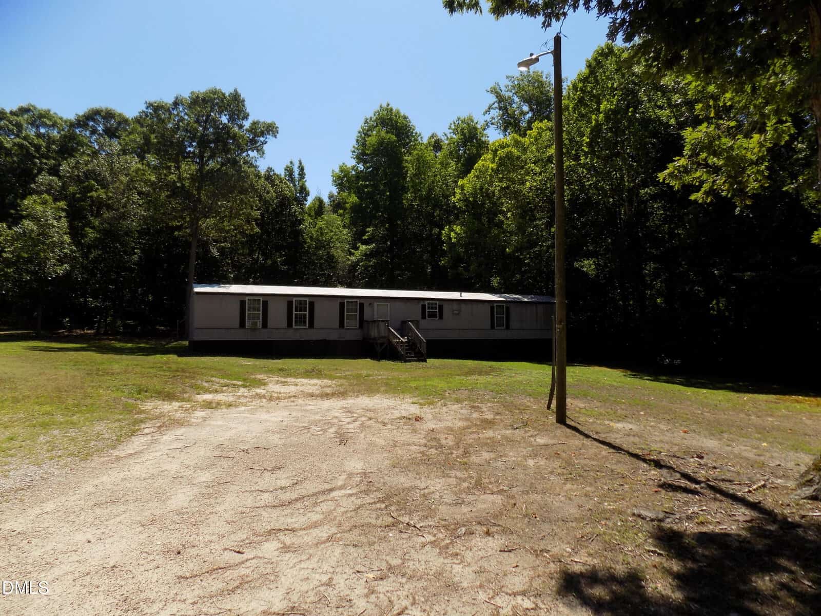 A single-story gray mobile home with front steps sits in a clearing surrounded by trees, under a clear blue sky. A dirt driveway and a wooden utility pole are visible in the foreground.