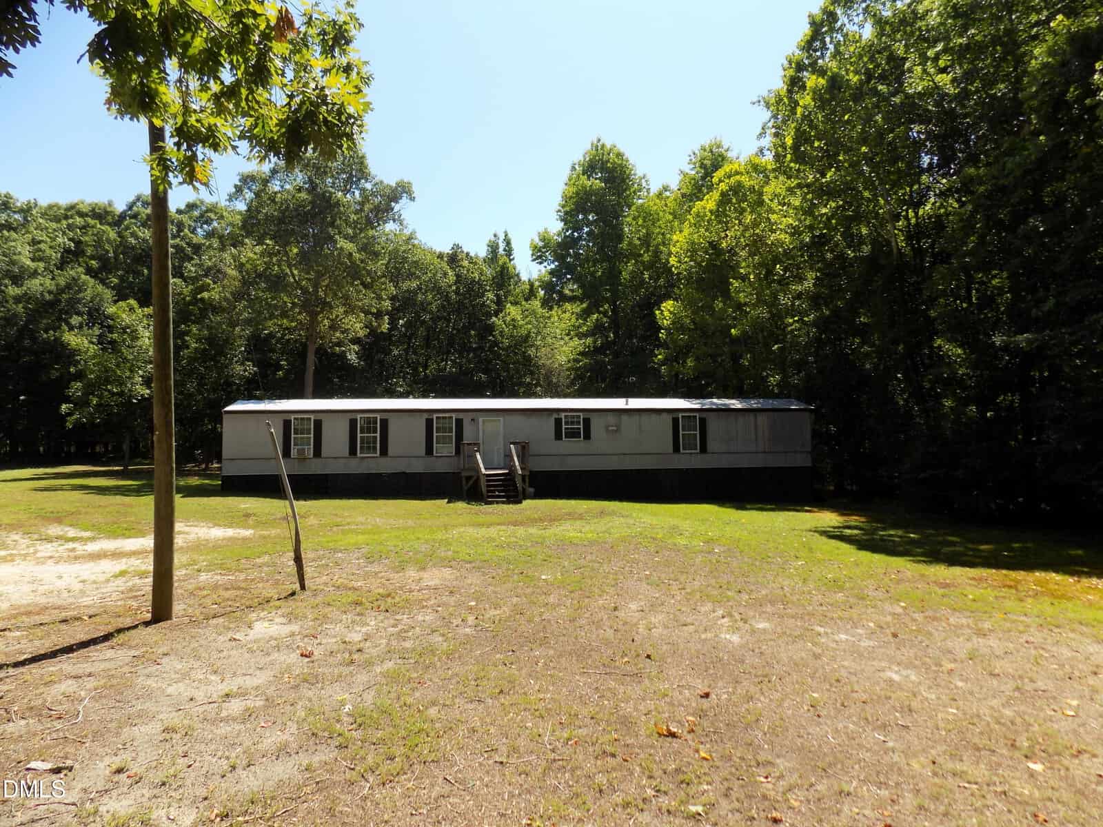 A long, single-story gray mobile home sits on a grassy, partially shaded lot at 0 E River Road in Louisburg, surrounded by tall green trees under a clear blue sky. Wooden steps lead to the front door.