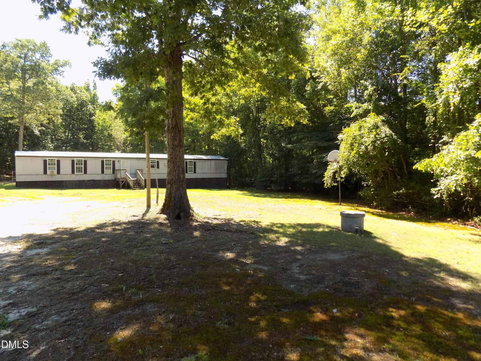 A single-story mobile home with steps leading to the entrance sits on a grassy lot surrounded by trees. A large tree and a basketball hoop are visible in the shaded front yard.