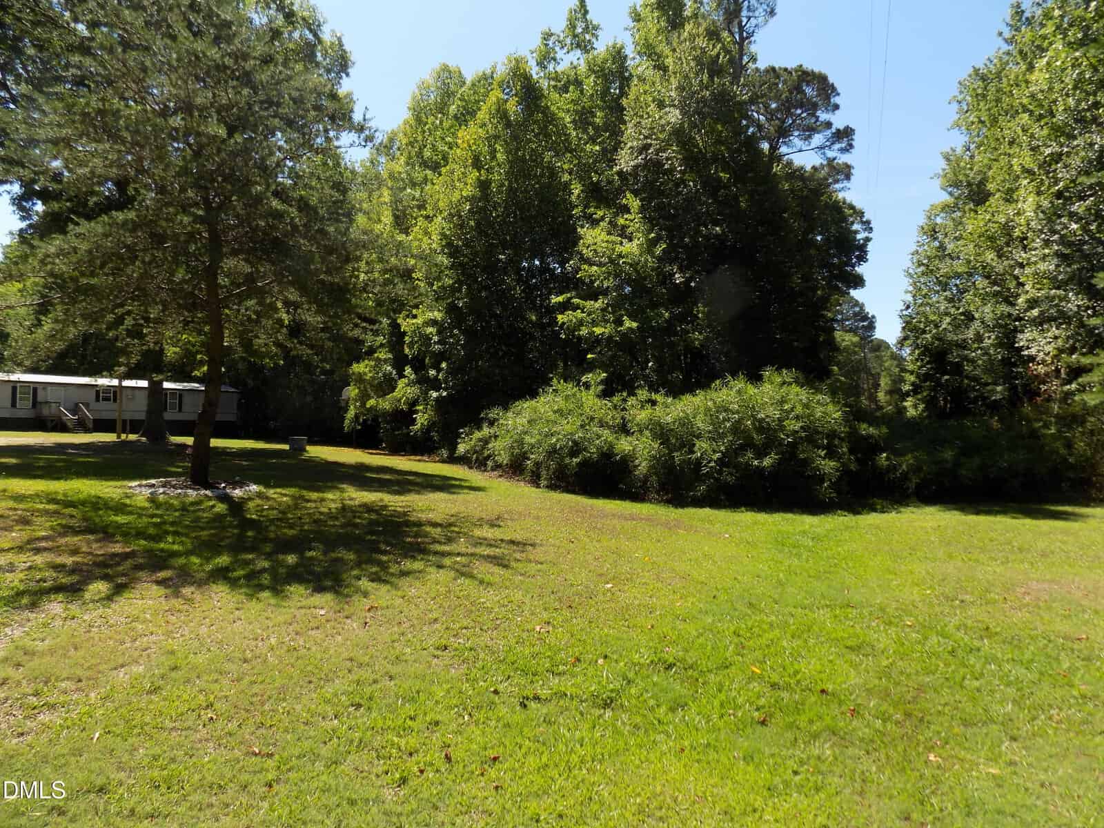 A grassy yard with scattered trees and bushes, bordering a wooded area. A white mobile home is partially visible on the left side under the trees, and the sky is clear and blue.
