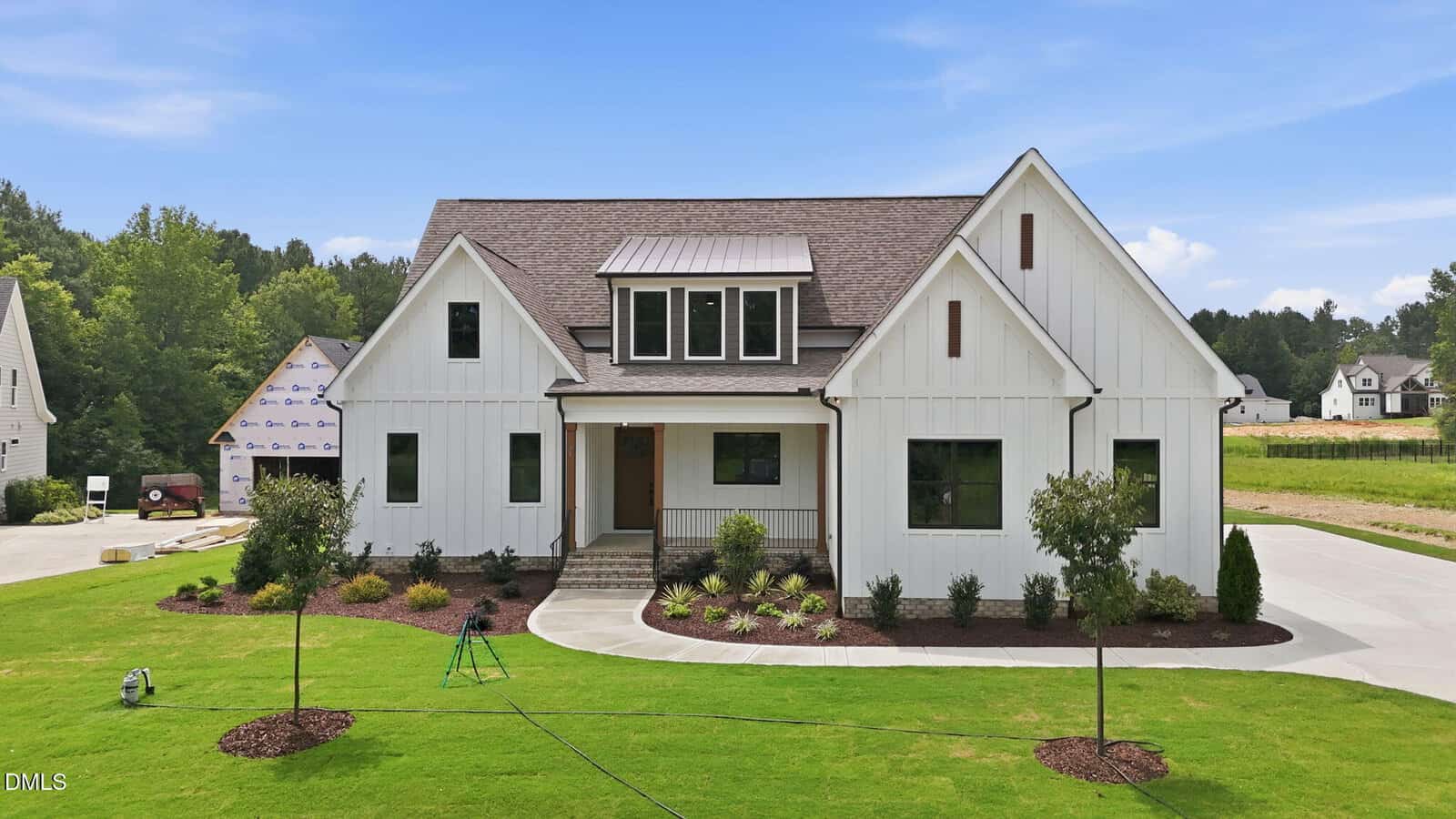 A modern white farmhouse with a gable roof sits on a well-manicured lawn. Young trees and shrubs are planted in mulched beds. A concrete driveway leads to the side of the house. Another home is partially visible nearby.
