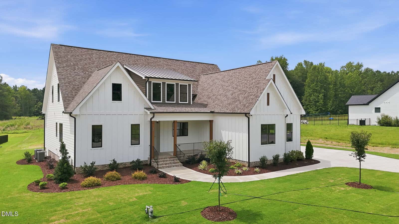 A two-story white house with a gable roof, covered front porch, and landscaped yard sits on a large green lawn with a driveway, surrounded by trees and another house in the background under a blue sky.