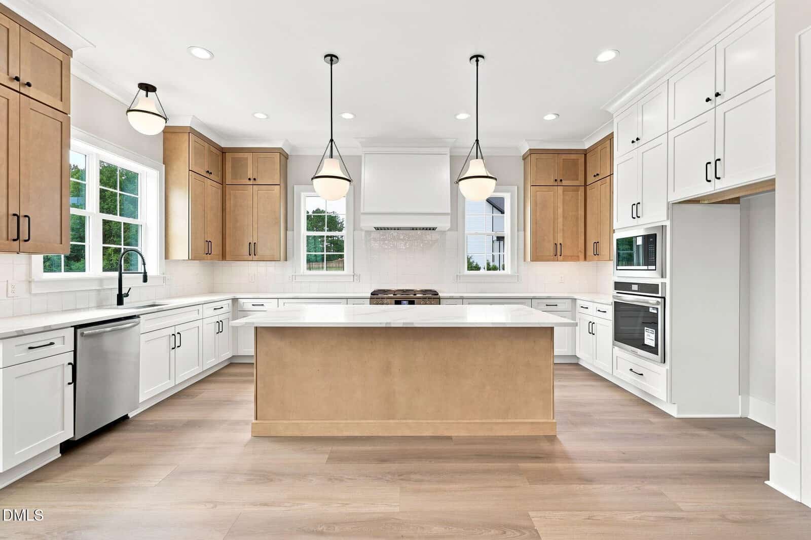 Modern kitchen with light wood and white cabinets, stainless steel appliances, a large island with a light wood finish, white countertops, three pendant lights, and large windows providing natural light.