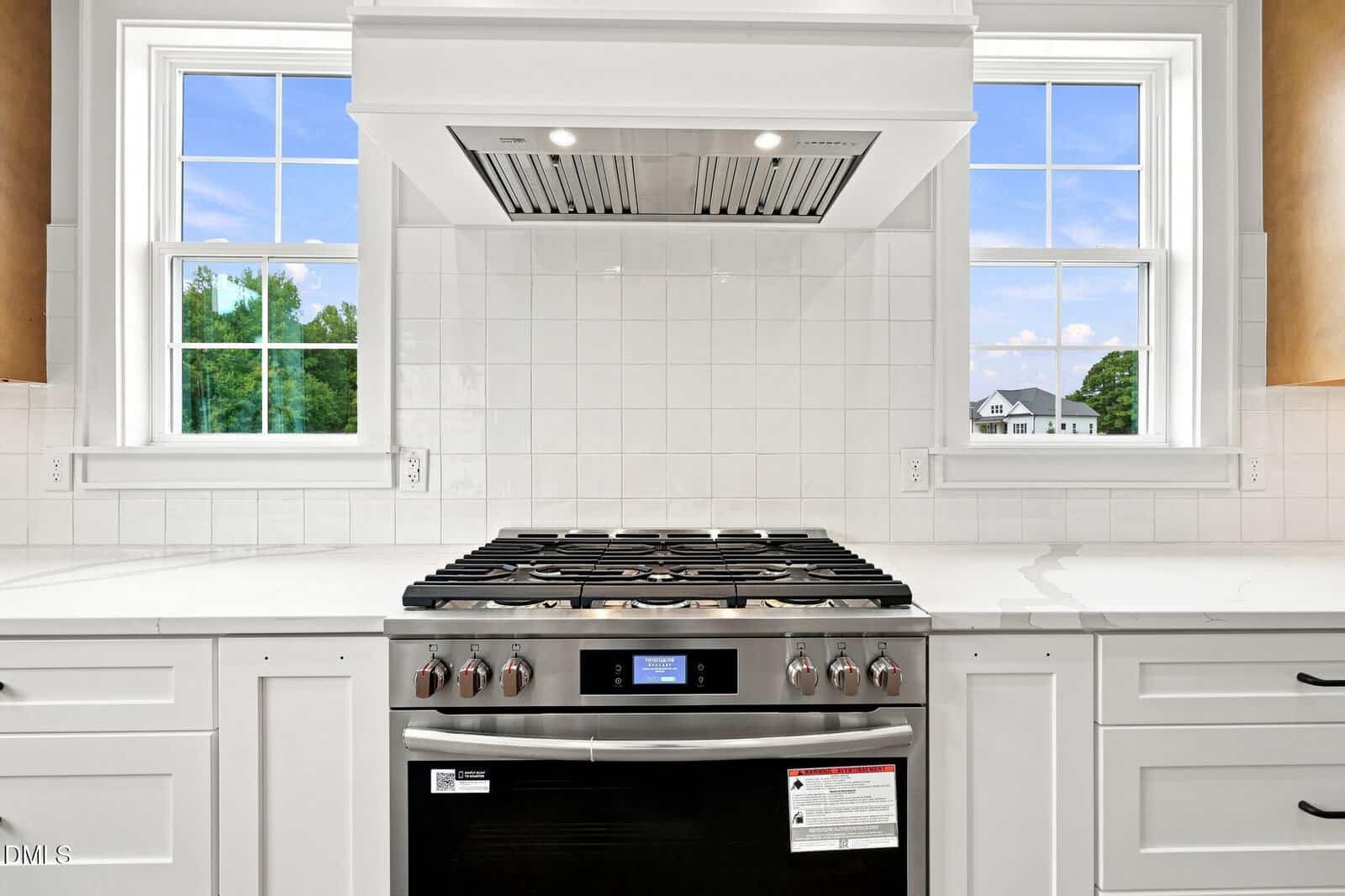 Modern kitchen with a stainless steel gas stove and built-in oven, white tiled backsplash, white cabinets, and two large windows letting in natural light.