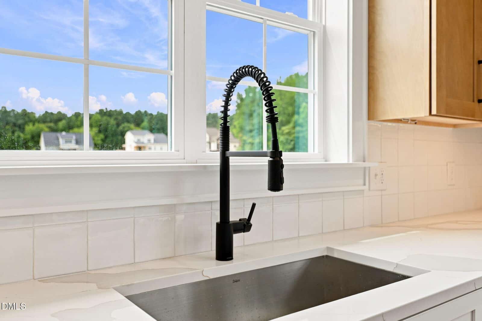 Modern black kitchen faucet with a flexible spring neck above a stainless steel sink, set in a white countertop, in front of a window showing a scenic outdoor view.