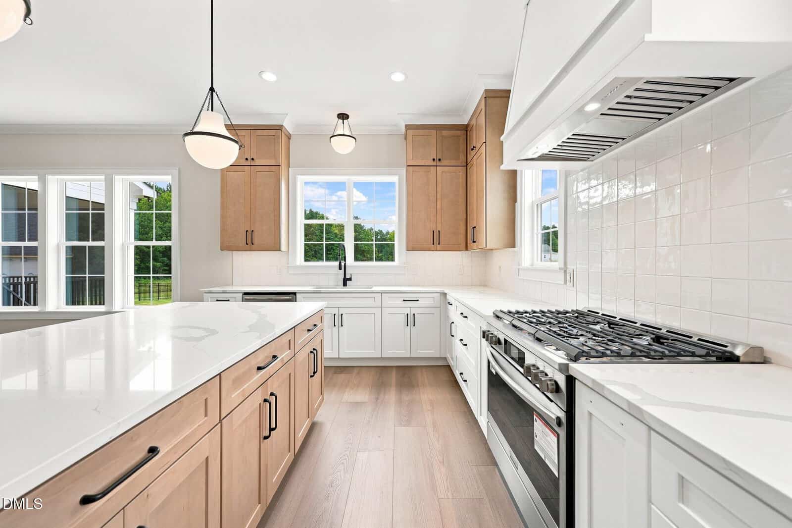 Modern kitchen with light wood cabinets, white countertops, a gas stove, white tile backsplash, black fixtures, pendant lights, and large windows overlooking a green yard.