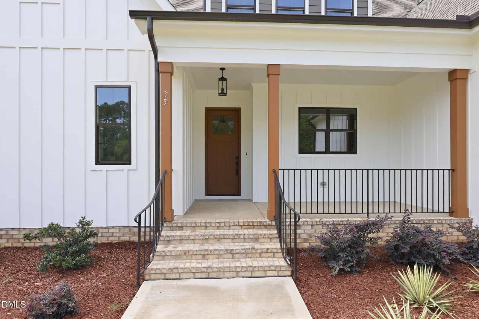 The image shows the front porch of a modern house with white siding, brown columns, a brown front door, a black railing, and landscaped mulch with shrubs along the walkway and steps leading to the entrance.