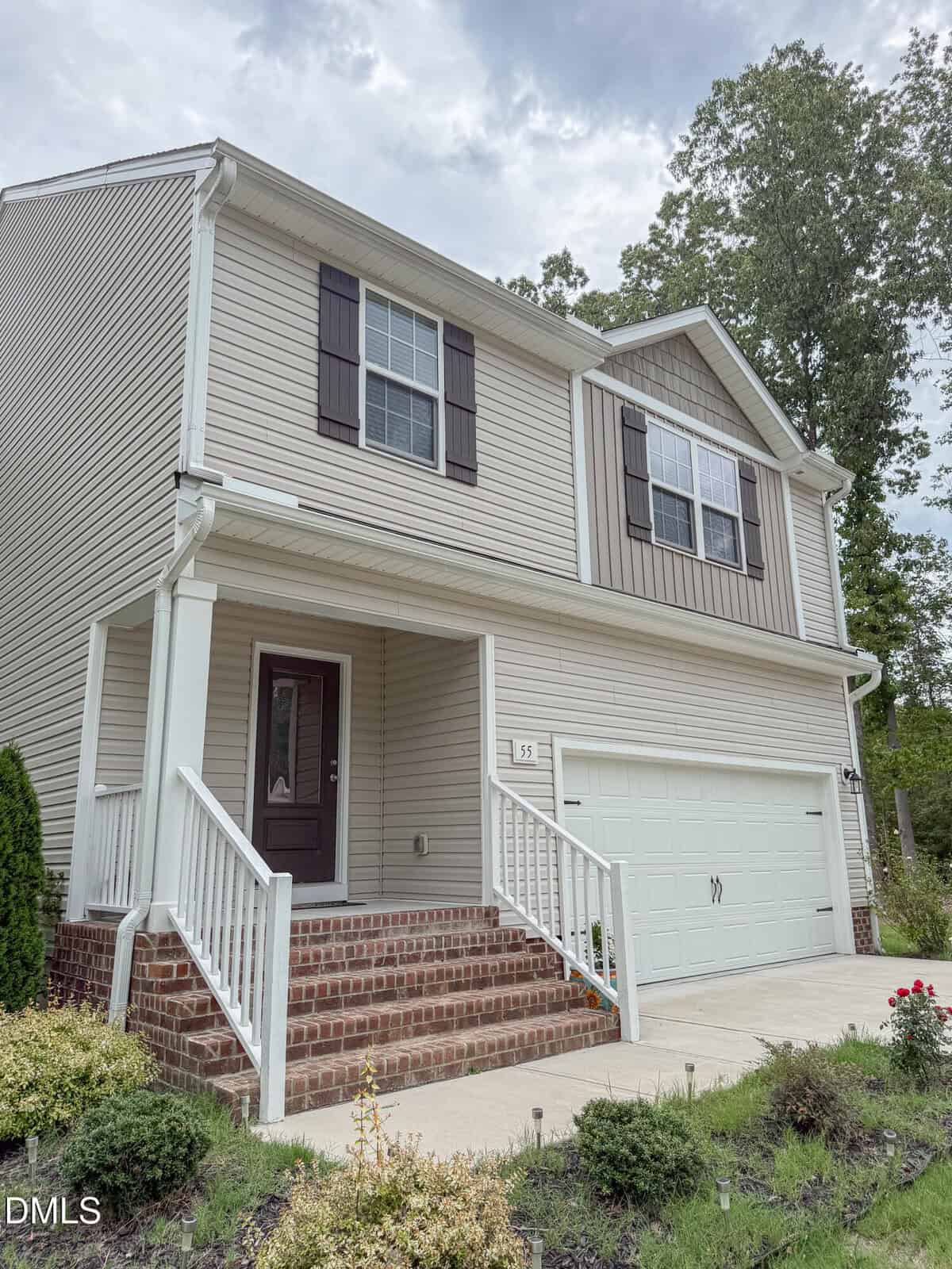 Two-story beige house with white trim, dark shutters, and a front porch with brick steps. The house has a double garage and is surrounded by greenery and small shrubs. The sky is cloudy.