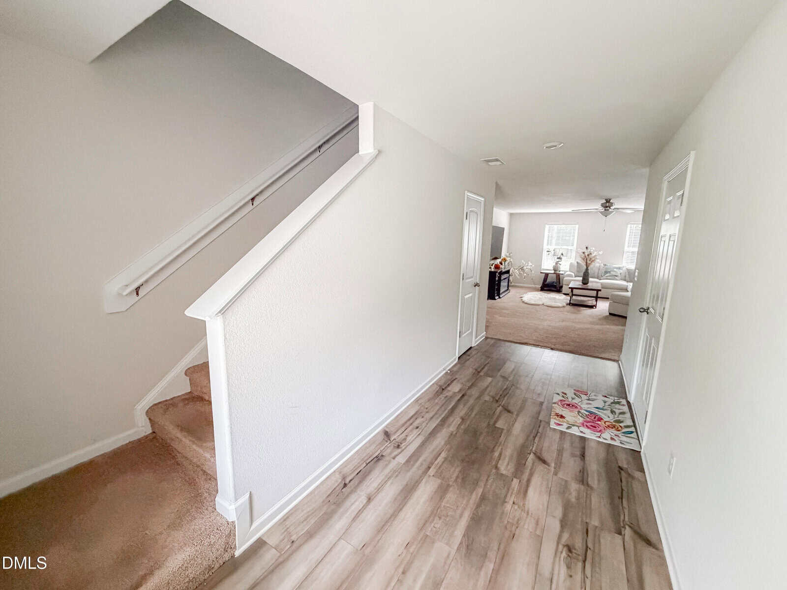 Bright hallway with light wood flooring, leading to a spacious living room. Carpeted stairs are on the left, and a white door is on the right. The living room has sofas, windows, and a ceiling fan.