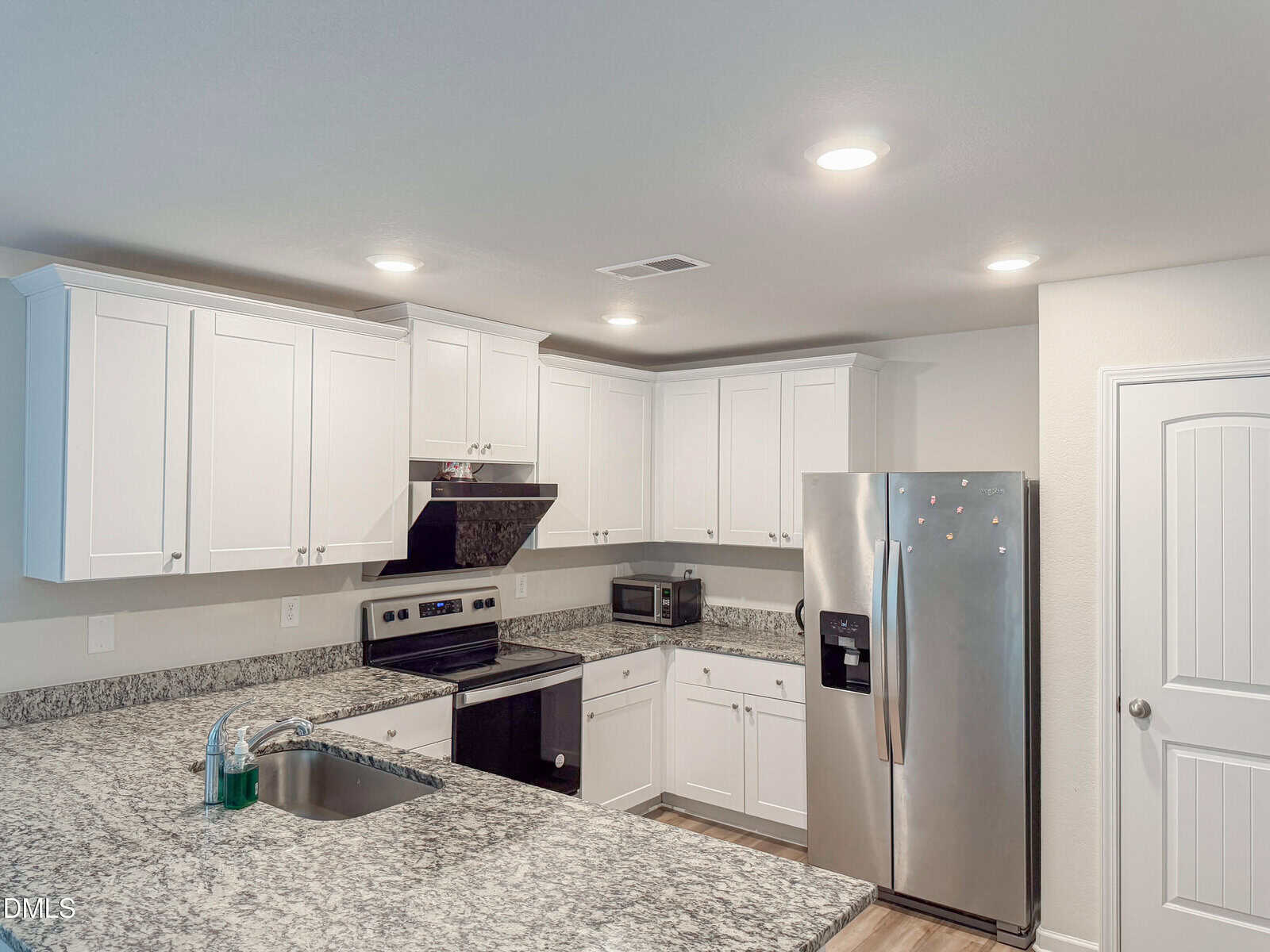 Modern kitchen with white cabinets, stainless steel appliances, granite countertops, a double-basin sink with soap and sponge, an electric stove with a black range hood, and a closed white door on the right.