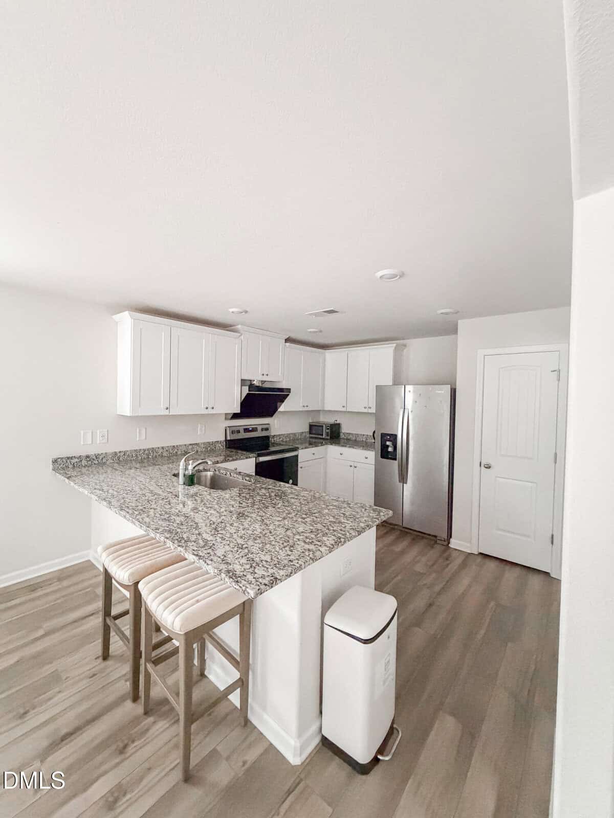 Modern kitchen with white cabinets, granite countertops, stainless steel appliances, a center island with two striped stools, wood-style flooring, and a white trash bin by the island. Walls and ceiling are white.