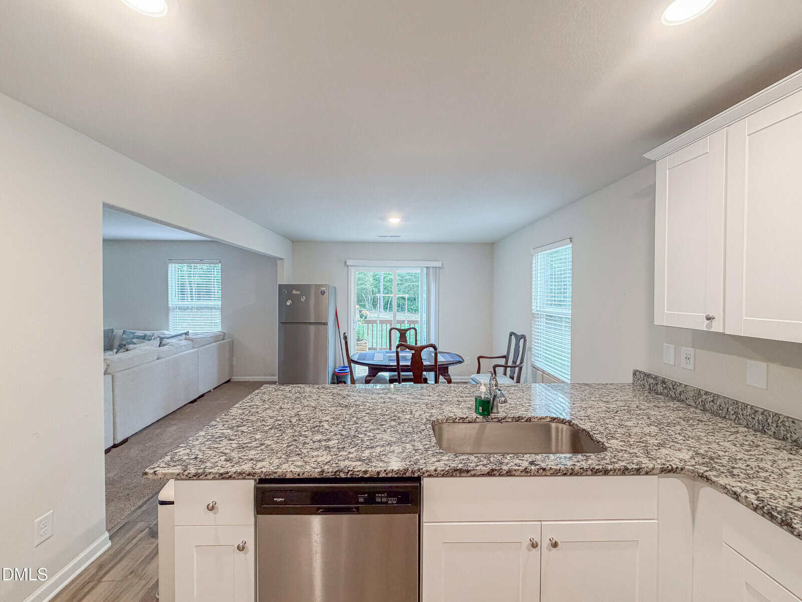 View from a granite kitchen island with a sink and dishwasher, overlooking a dining area with a table and chairs, a refrigerator, and an adjacent living room with a sectional sofa. Large windows let in natural light.