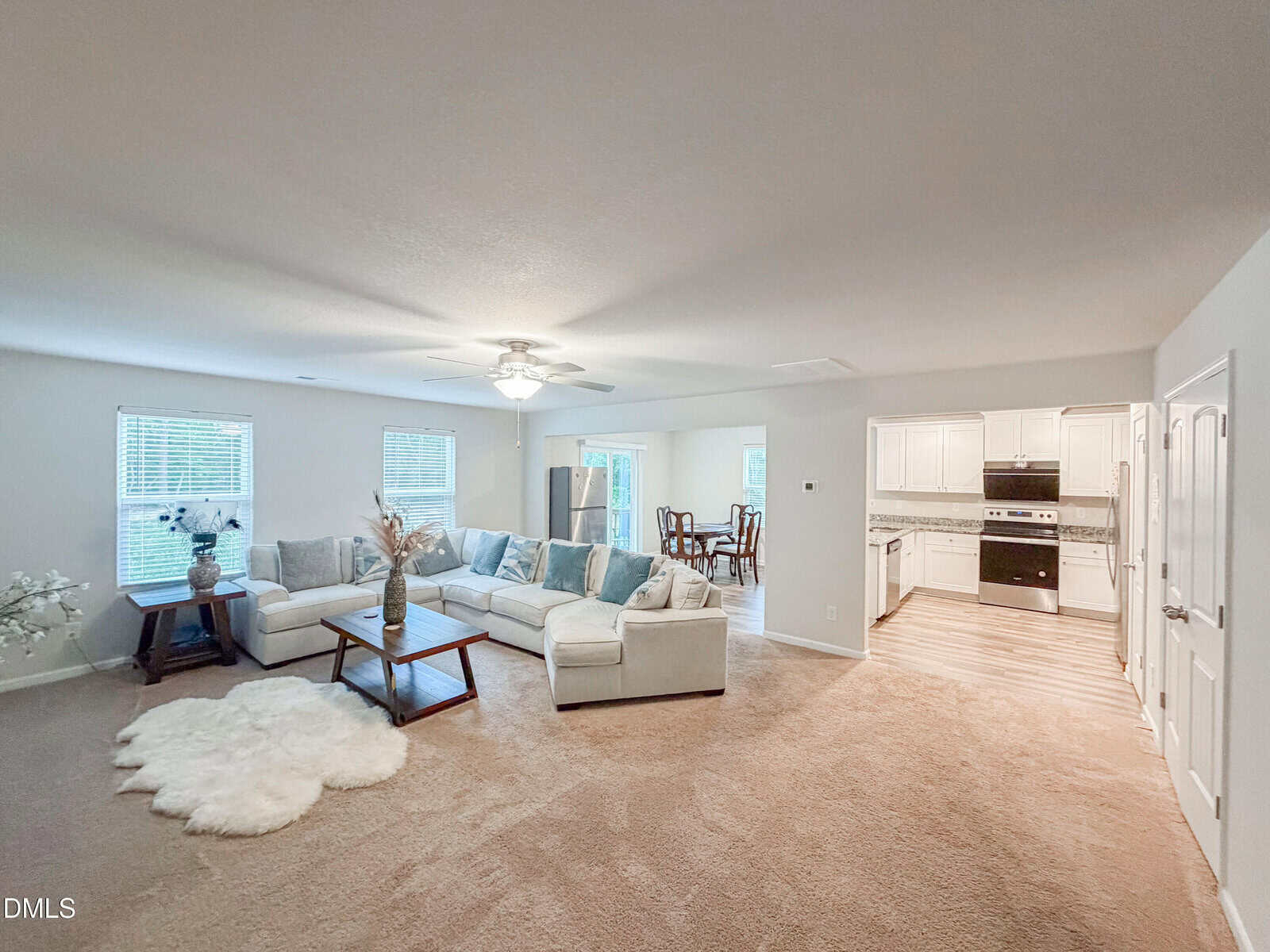 Spacious living room with beige carpet, a white sectional sofa, a throw rug, coffee tables, large windows, and an open view into a dining area and kitchen with white cabinets.