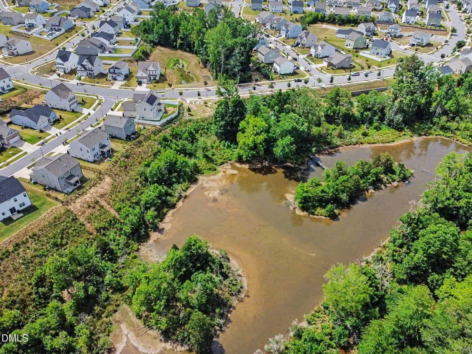 Aerial view of a suburban neighborhood with rows of houses surrounding a small pond and wooded area, separated by a road and greenery.