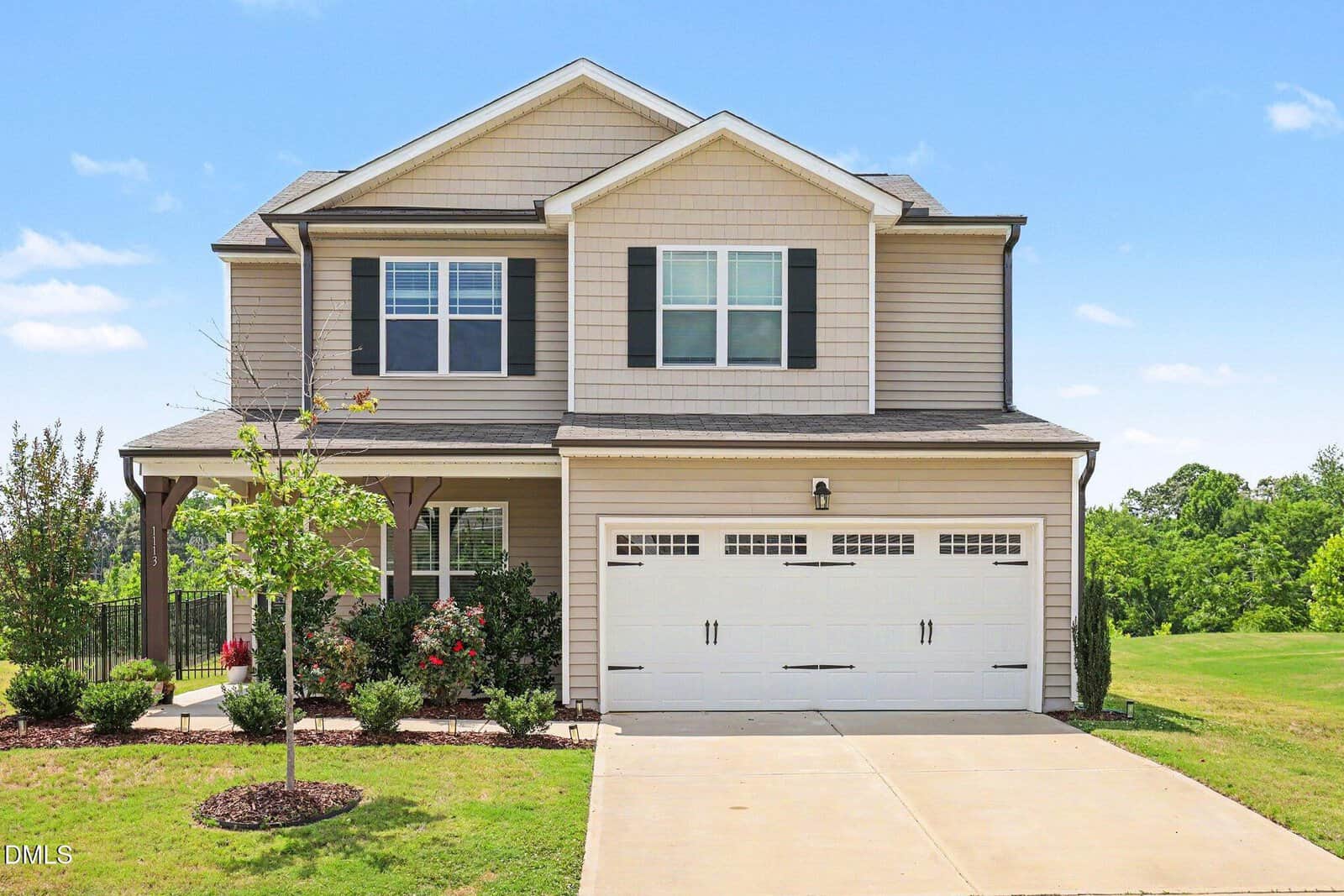 A two-story beige house at 1113 Shadow Shade Drive in Wake Forest features a double garage, black shutters, covered porch, and small front yard with young trees and shrubs, set against green grass and trees under a blue sky.