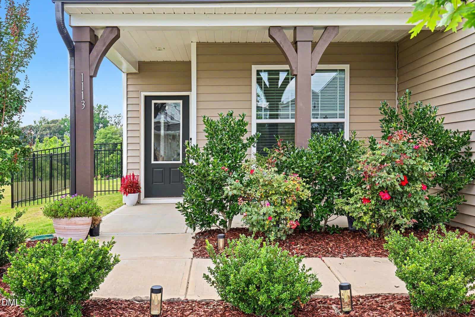 A beige house with a covered porch features a black door, large window, wooden pillars, and vibrant green shrubs and flowering plants lining the walkway. A potted plant sits near the entrance.