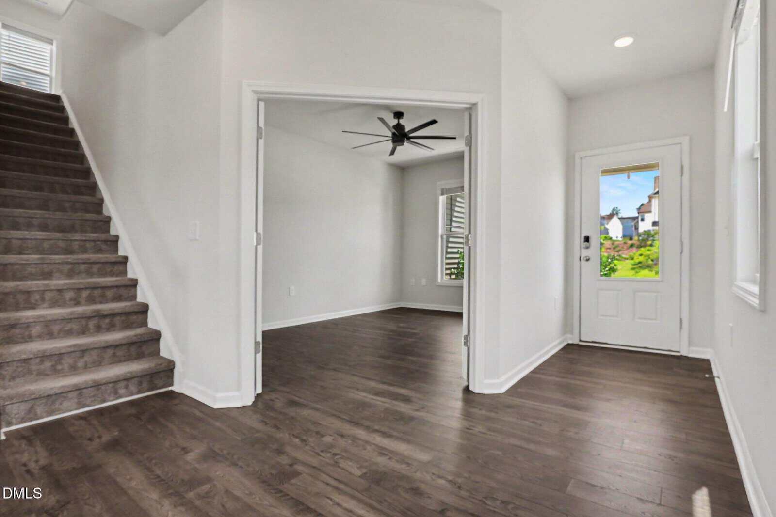 Interior view of a modern home with dark wood floors, white walls, a staircase on the left, a room with double doors and a ceiling fan in the center, and a front door with a window on the right. Natural light fills the space.