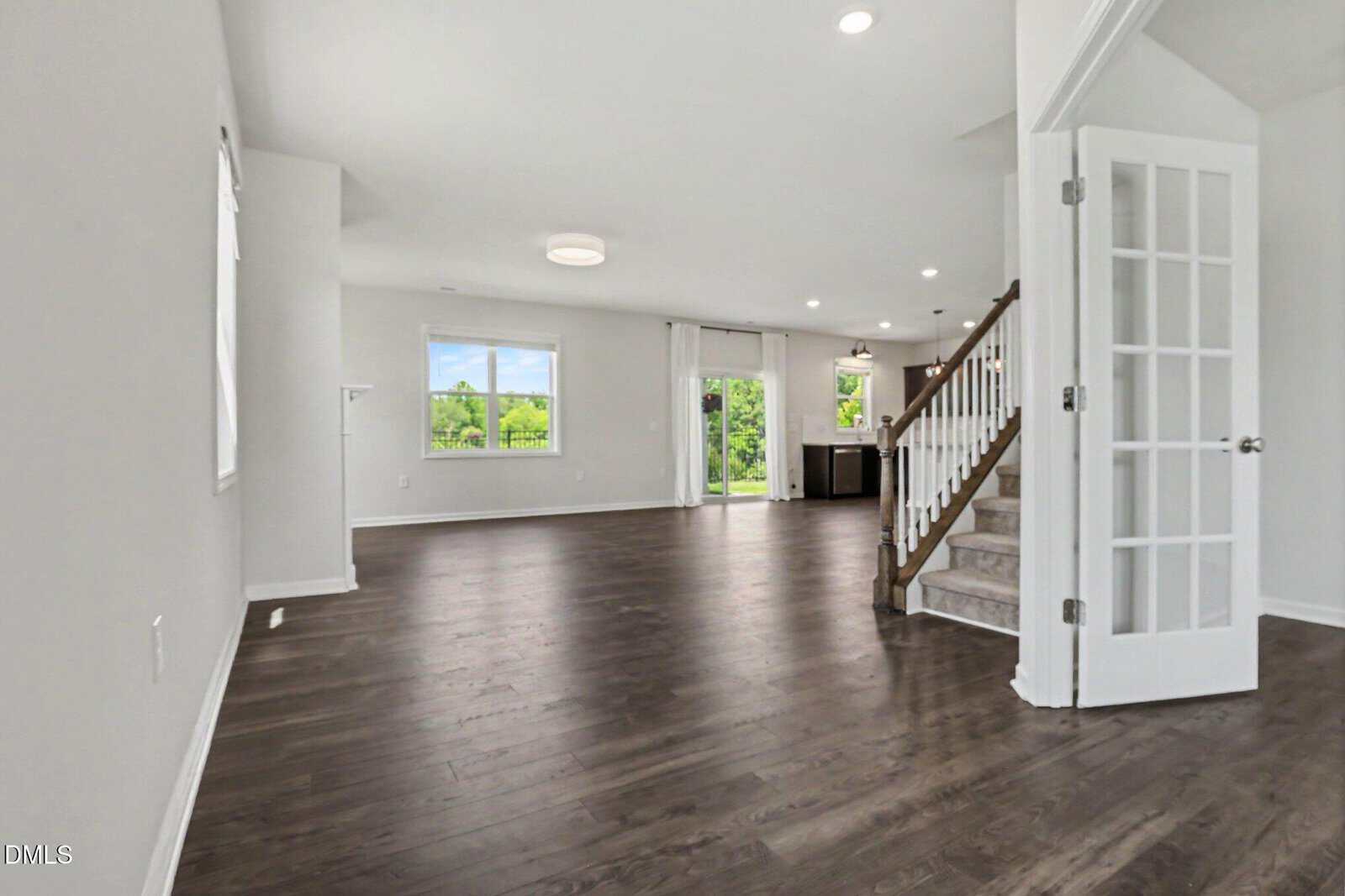 Open-plan living area with dark wood floors, white walls, large windows, glass doors to the outside, and a staircase with white railings. French doors lead to a separate room on the right. Bright and modern interior.