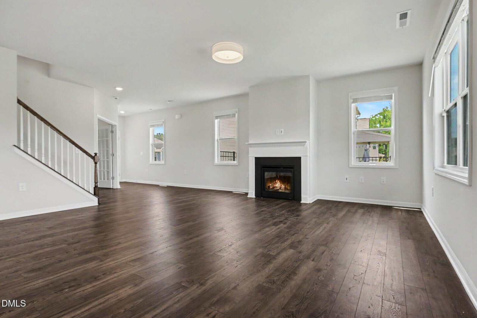 A spacious, empty living room with dark wood floors, white walls, a modern fireplace, large windows, and a staircase with a white railing leading upstairs.