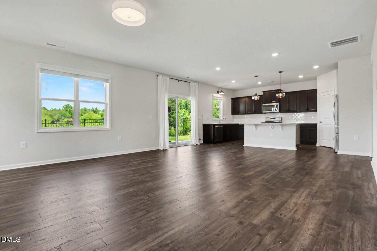 Spacious open-concept living area with dark wood floors, large windows, sliding glass doors, and a modern kitchen with dark cabinets, white island, and stainless steel appliances.