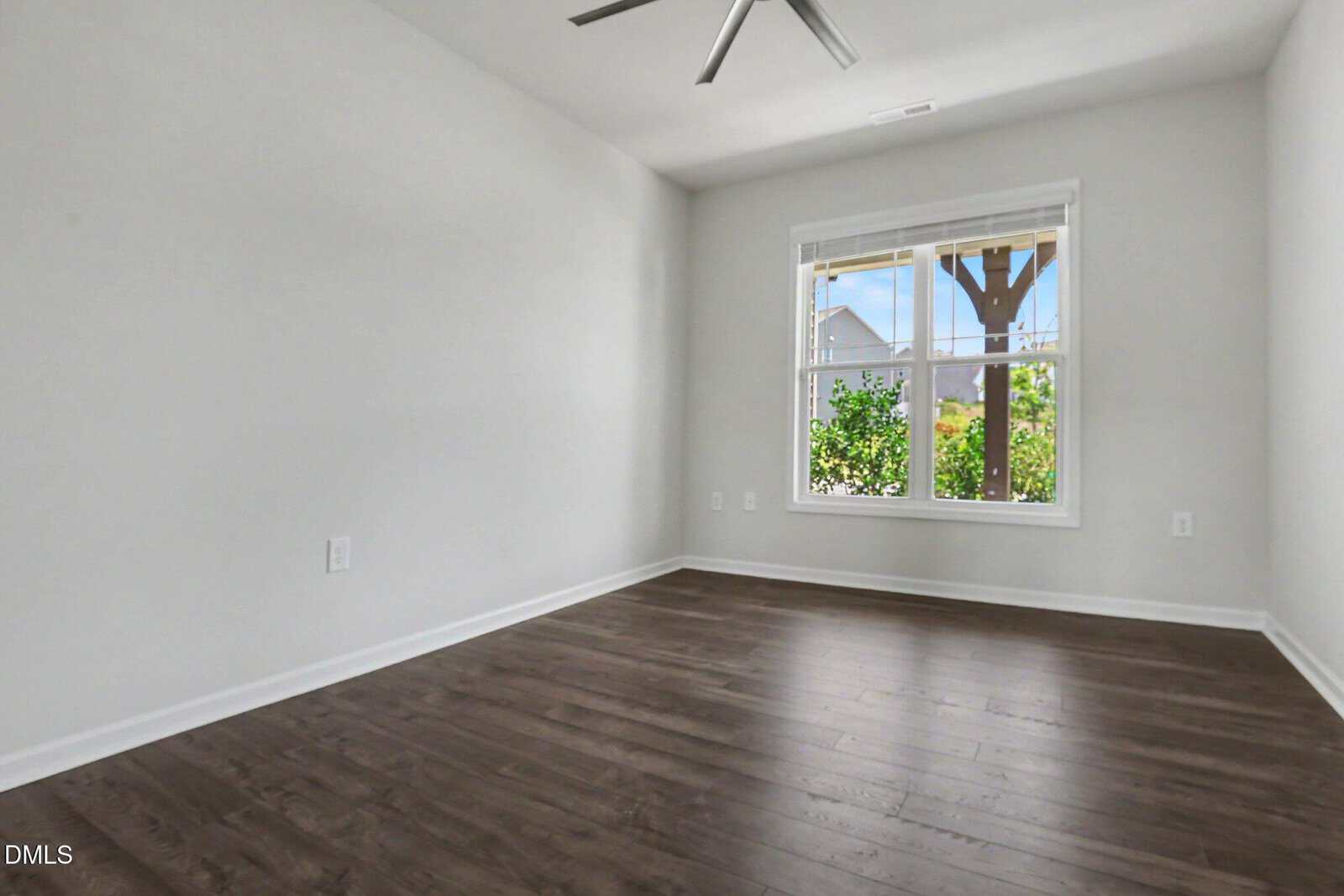 A bright, empty room with light gray walls, dark wood flooring, a large window with white trim, and a ceiling fan. Greenery and a pergola are visible outside the window.