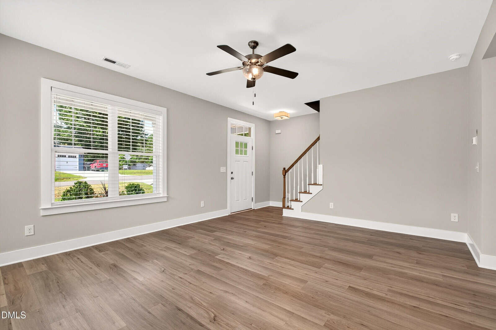 A bright, empty living room with light gray walls, wood flooring, a ceiling fan with light, a large front window, and a white door near a staircase with a wooden handrail.