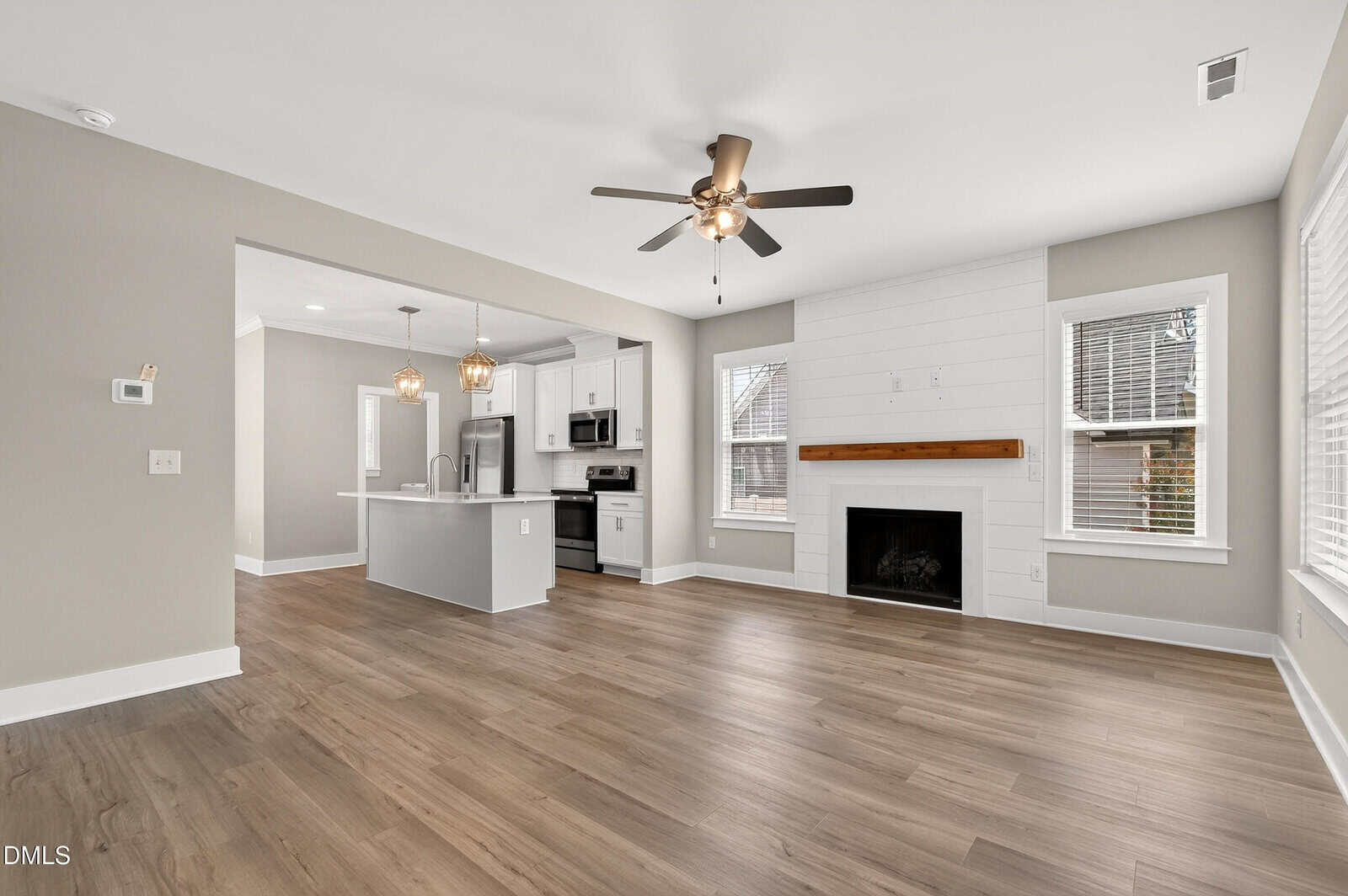 Open-concept living area with wood floors, a ceiling fan, a white shiplap fireplace, and large windows. The modern kitchen features stainless steel appliances, a center island, and pendant lights. Neutral gray and white tones throughout.