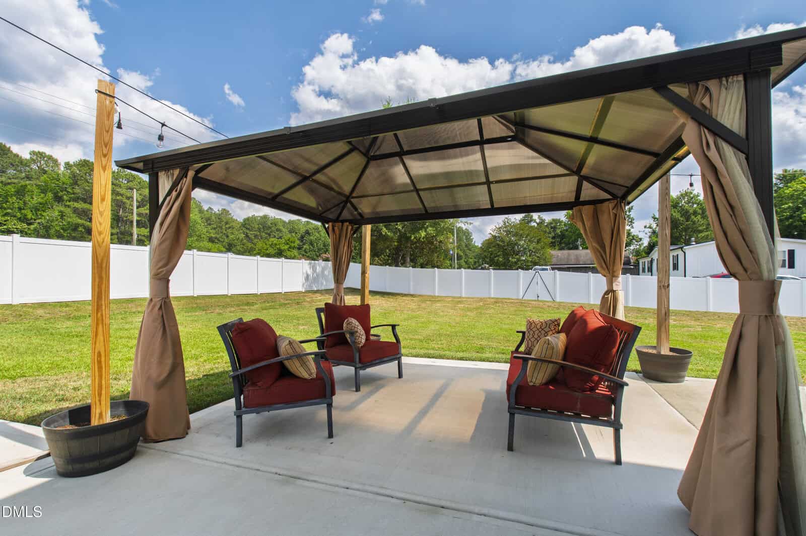 A backyard patio with a gazebo featuring a metal roof and beige curtains. Inside are two cushioned chairs and a loveseat with red cushions, all arranged on a concrete slab. The yard is grassy and surrounded by a white fence.