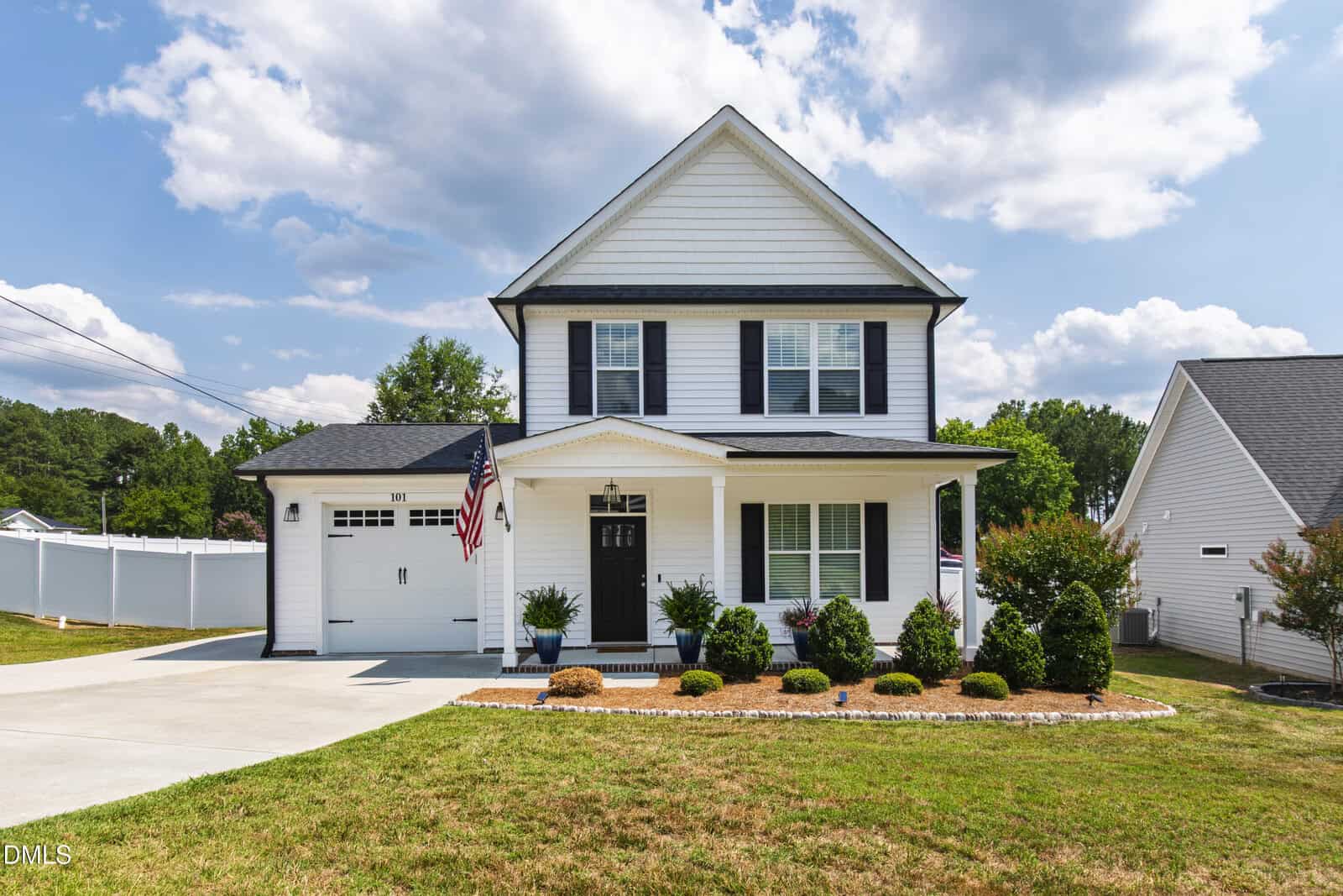 A two-story white house with black shutters at 101 Averly Court, Stem features a covered front porch, attached garage, and an American flag. The manicured lawn, neat bushes, and paved driveway complete the charming curb appeal under a partly cloudy sky.