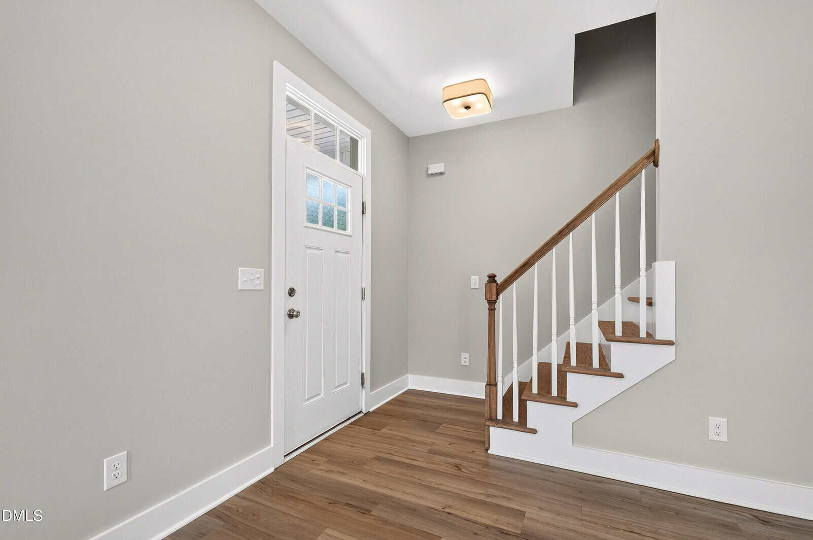Bright entryway with a white front door, side window panels, and a wooden staircase with white spindles. Light gray walls and hardwood floors create a clean, modern look. Ceiling light fixture above.