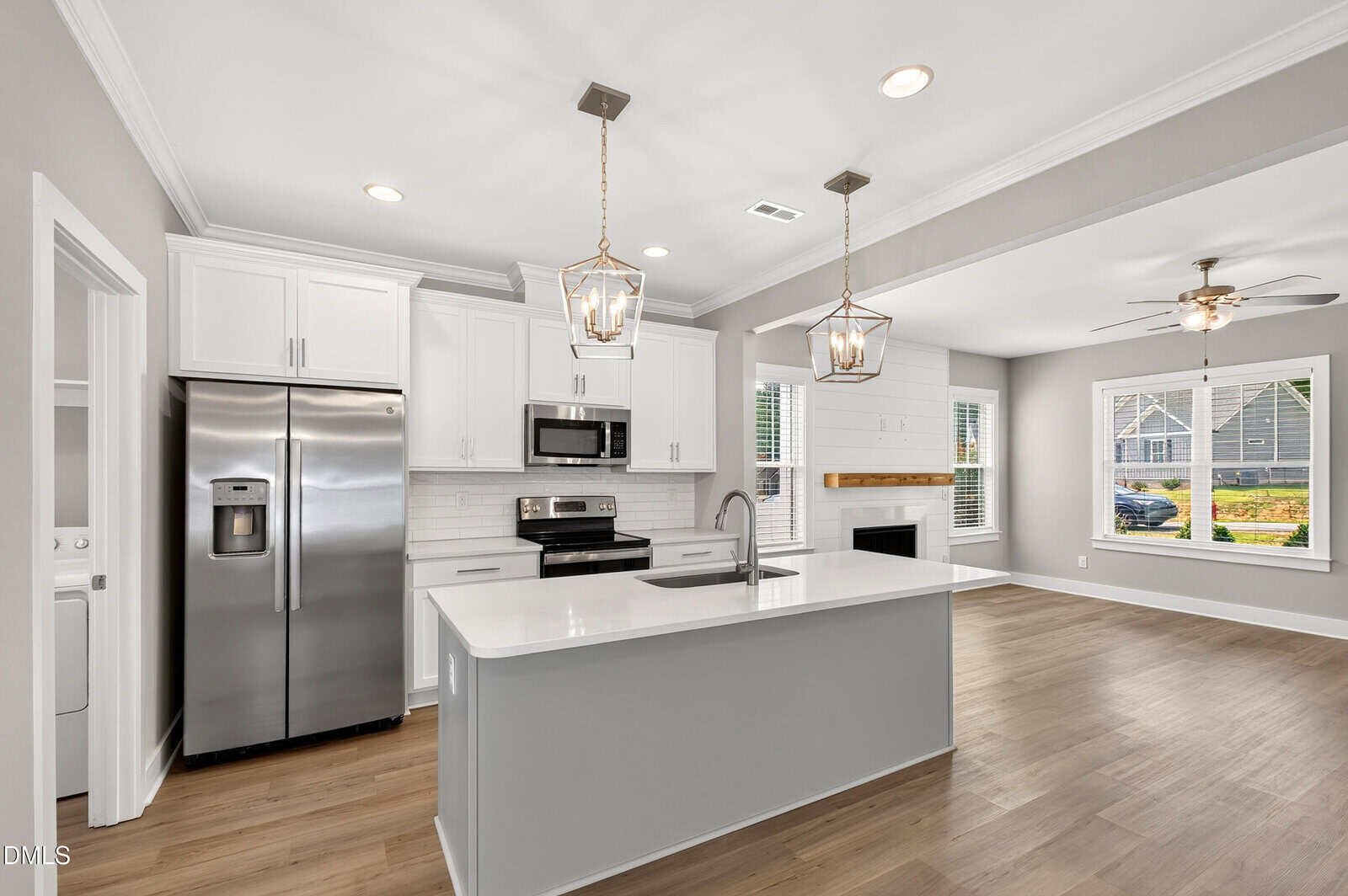 Modern kitchen with white cabinets, stainless steel appliances, a large island with a sink, two pendant lights, and an open floor plan leading to a living area with a fireplace and large windows.