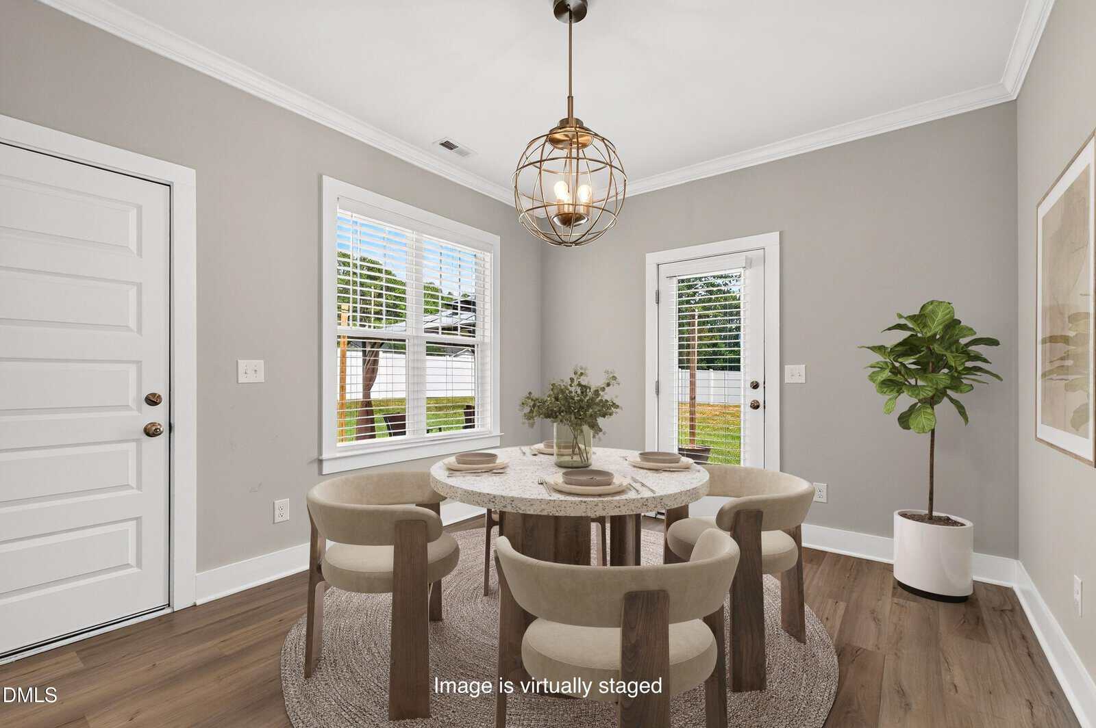 A virtual staging of a dining room with a round table set for four, beige chairs, a plant, and modern light fixture. Large windows and a glass door let in natural light.