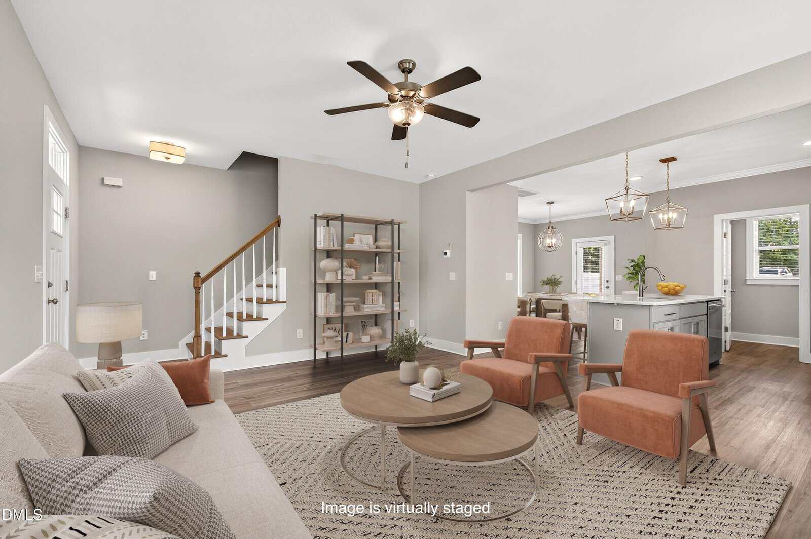A modern living room with neutral walls, a ceiling fan, a beige sofa, two orange chairs, a round coffee table, and a rug. The area opens to a kitchen with an island and pendant lights. The image is virtually staged.