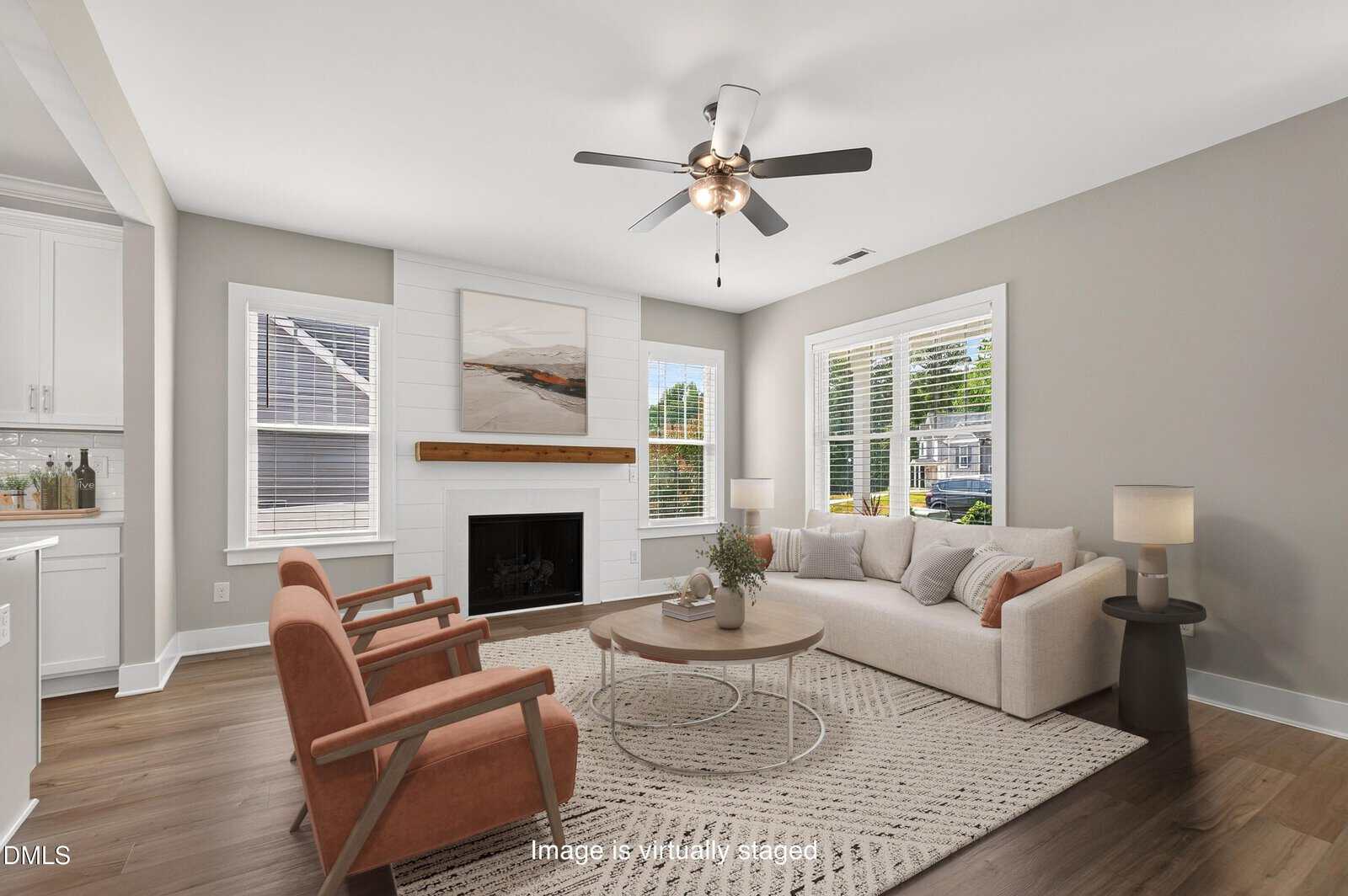 A living room at 101 Averly Court features a beige sofa, two orange chairs, a round coffee table, fireplace, wall art, a rug, and large windows. Hardwood floors and neutral walls stem from a modern design sensibility, creating a bright and inviting feel. Text at the bottom notes the image is virtually staged.