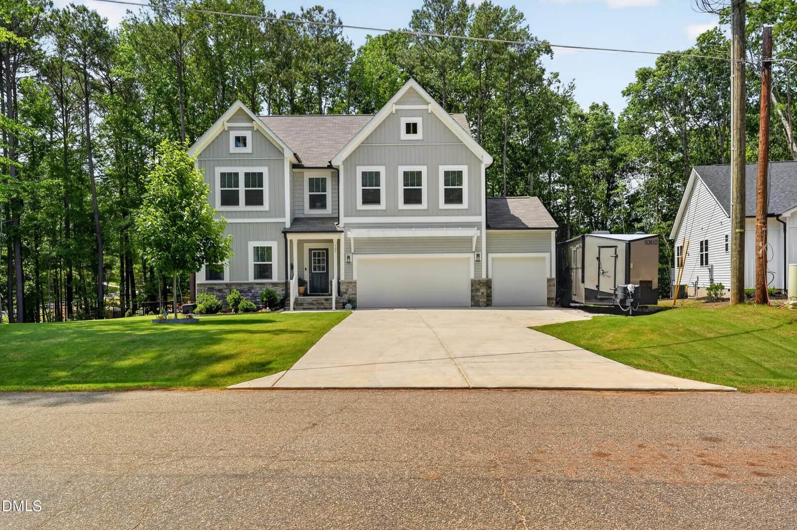 Beautiful two-story gray house with white trim and stone accents at 101 Yuma Drive, Louisburg. Features a three-car garage, large concrete driveway, green lawn, trees in the background, and a small shed to the right side.