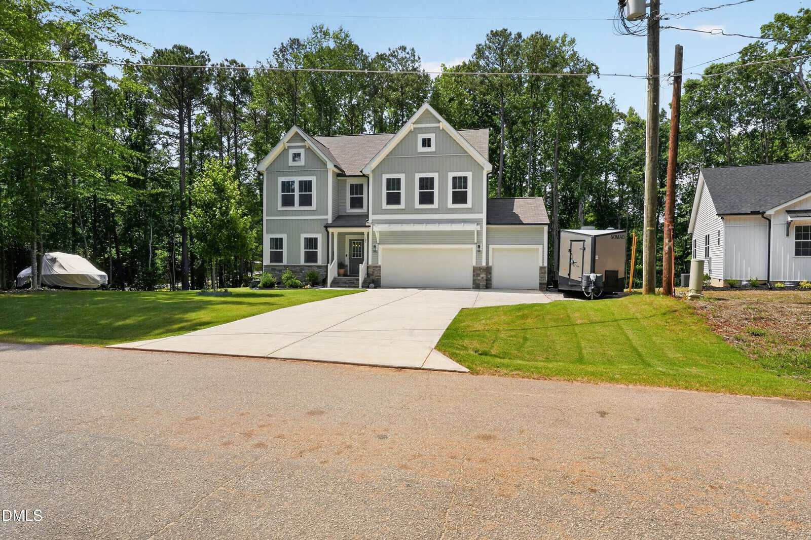 A two-story modern gray house with white trim, a three-car garage, and a wide driveway, surrounded by a green lawn and trees, viewed from the street on a sunny day.