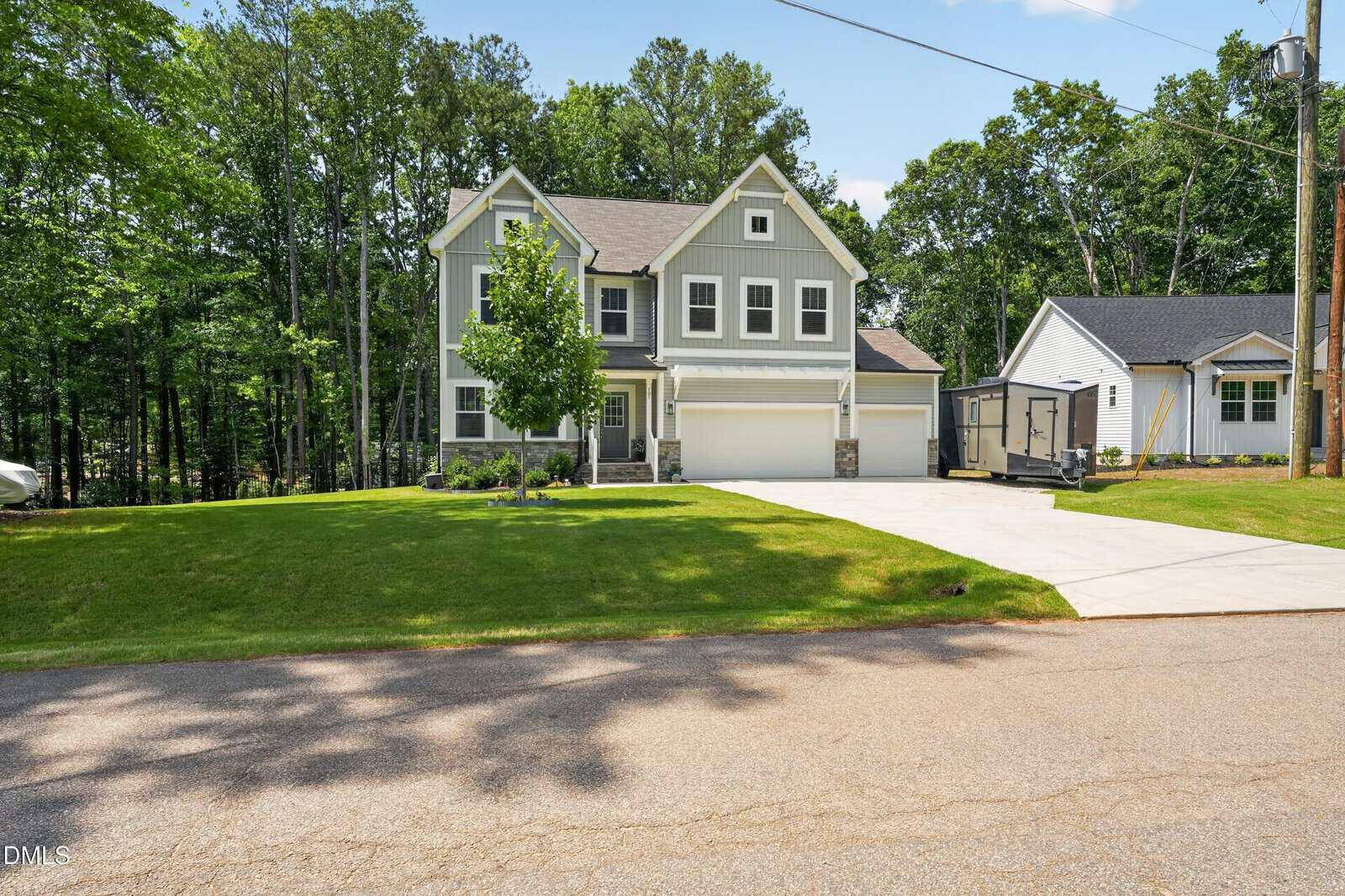 A two-story gray house with white trim, a double garage, and a well-kept lawn is shown on a sunny day, surrounded by tall trees. A small shed and a neighboring house are visible on the right.
