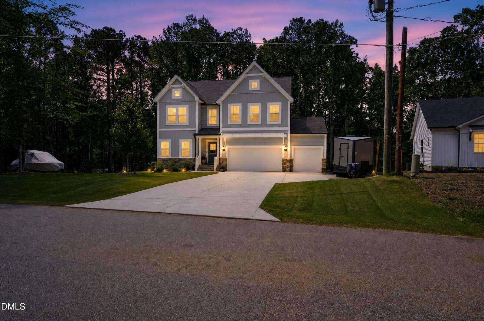 A two-story suburban house with lit windows, a three-car garage, and a spacious driveway at dusk, surrounded by trees and neighboring homes under a purple and blue sky.