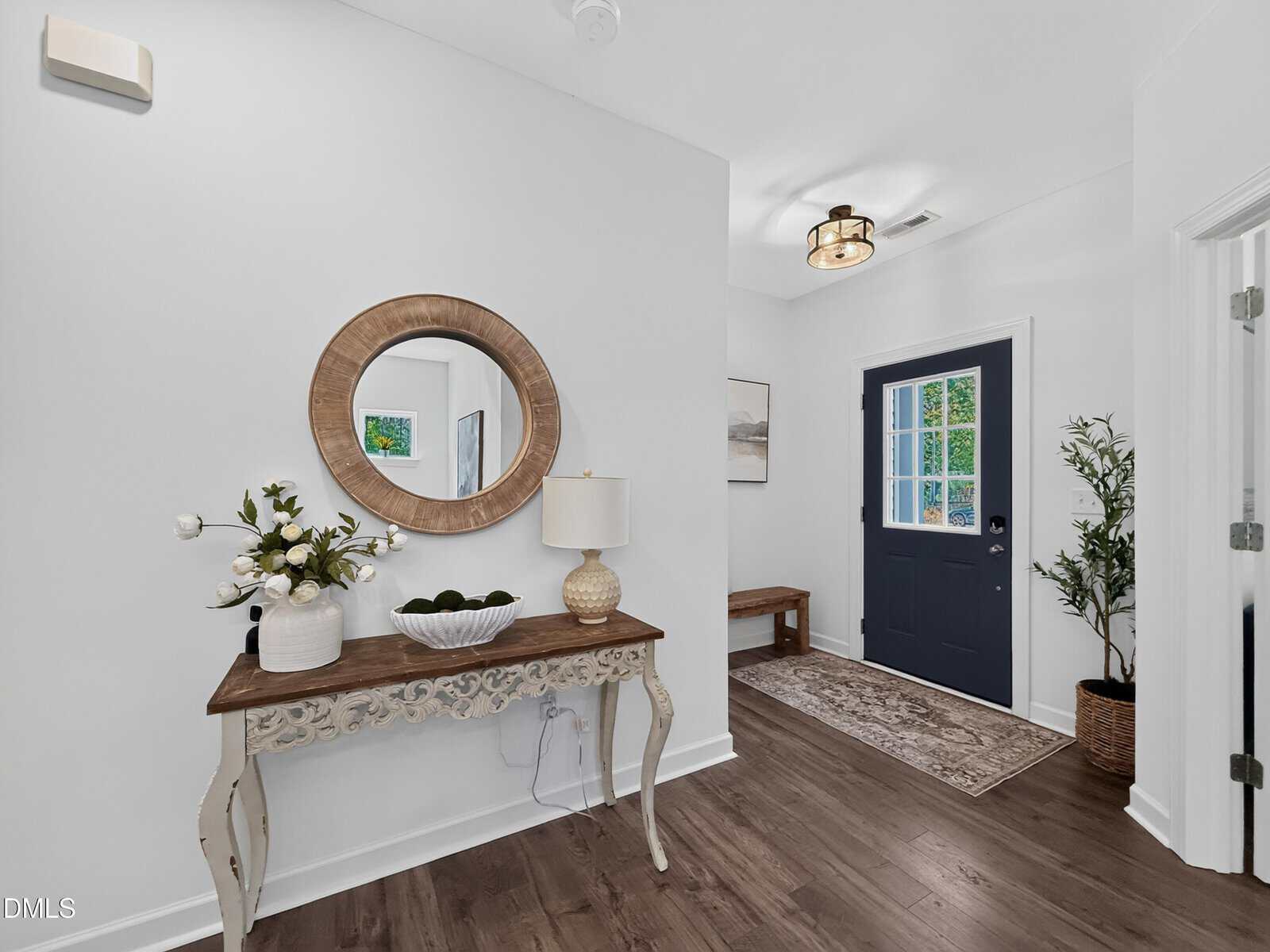 A bright entryway with a dark door, decorative console table, mirror, lamp, vase of flowers, bench, rug, and potted plant on dark wood flooring against white walls.