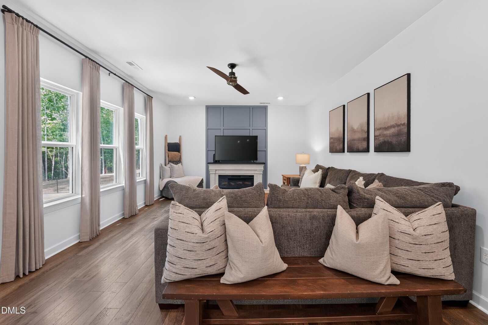 Modern living room with a gray sectional sofa, neutral pillows, a wooden bench, TV above a fireplace, large windows with drapes, wood floors, and two landscape art pieces on the wall.
