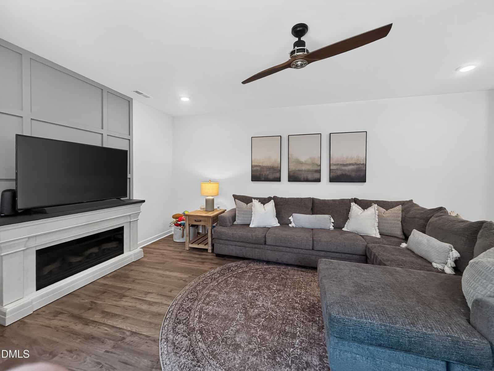 Modern living room with a gray sectional sofa, round brown rug, TV on a white fireplace, art on the wall, wooden side table with a lamp, and a ceiling fan. The room has wood flooring and neutral decor.